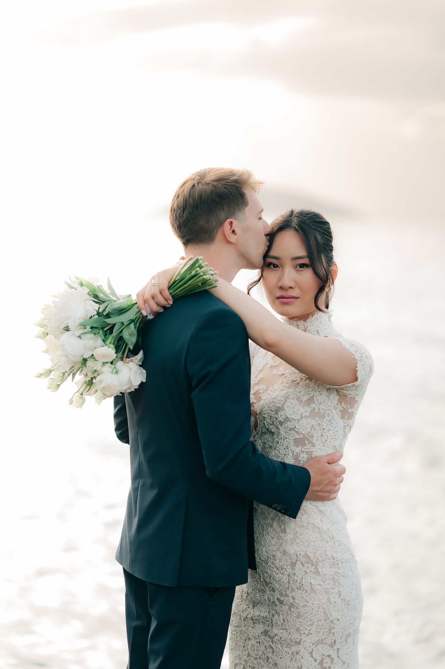 Groom kissing bride's cheek on sunlit beach, bride in lace mermaid dress holding large white tulip bouquet.