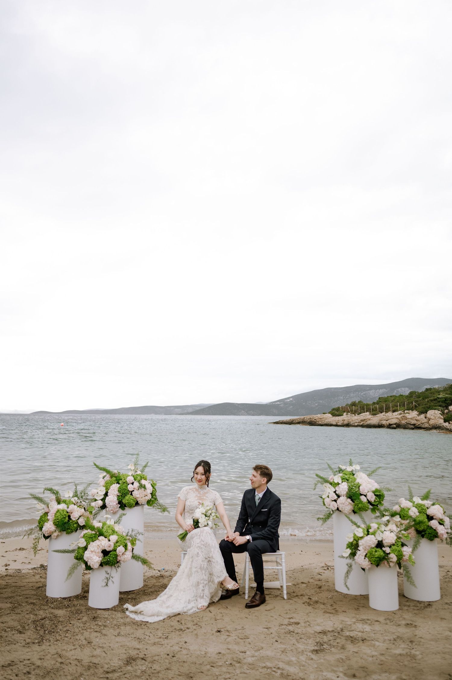 Bride and groom sit together on beach ceremony chairs surrounded by elegant white floral arrangements.