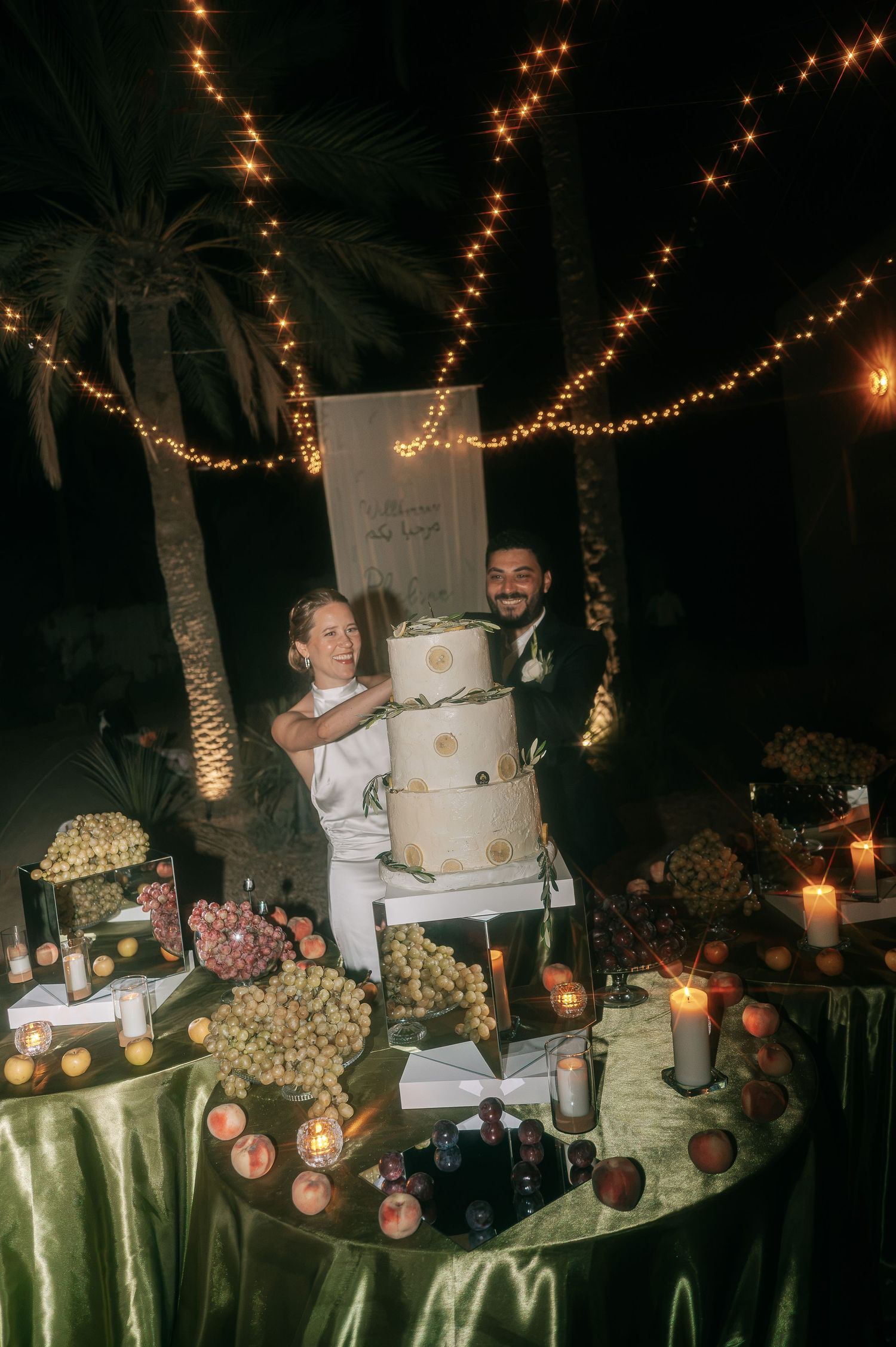 Bride and groom cut tiered wedding cake at dessert table adorned with fruit and candles.