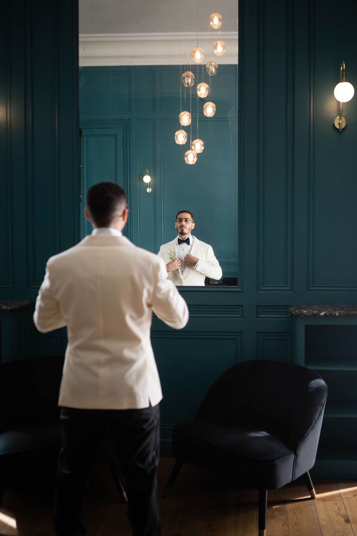 Groom in white tuxedo jacket adjusting bow tie in mirror of teal room.
