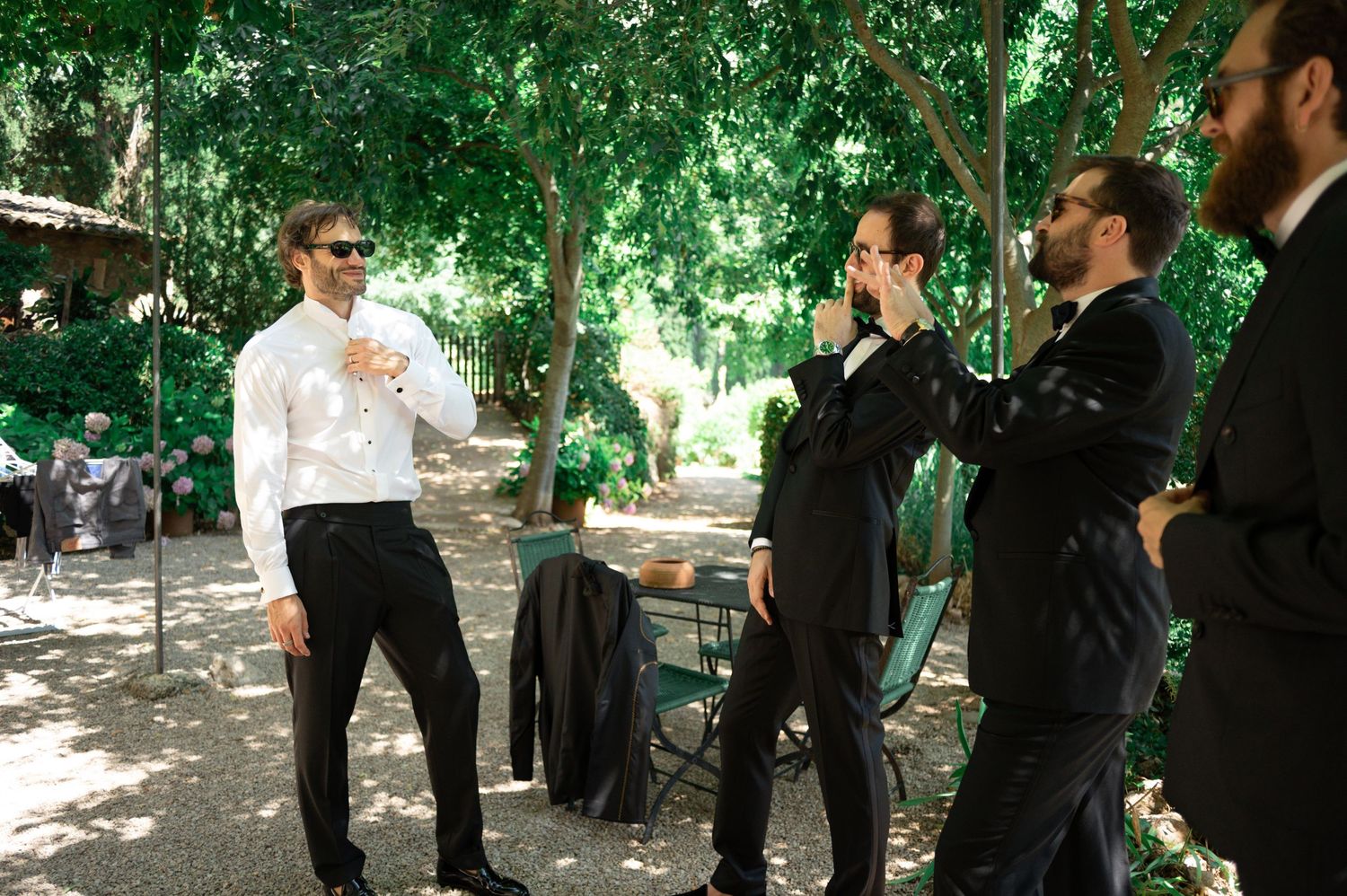 Groomsmen in black suits and white shirts socializing outdoors in a shaded garden setting.