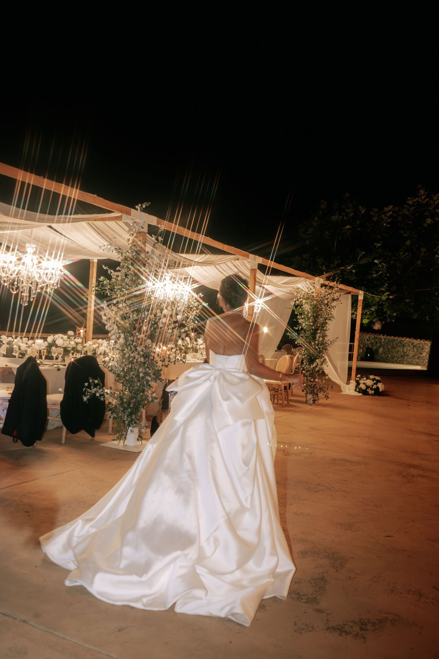 Bride in stunning white ballgown with large bow twirls near chandelier-lit outdoor wedding reception tent.