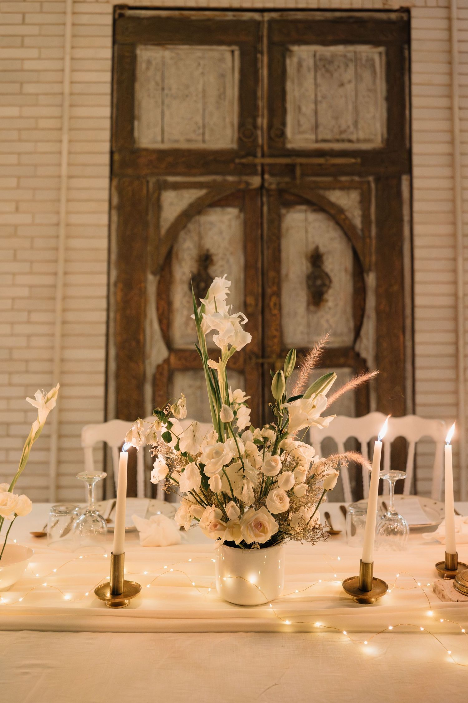 White floral centerpiece with candles and fairy lights on a wedding table before rustic doors.