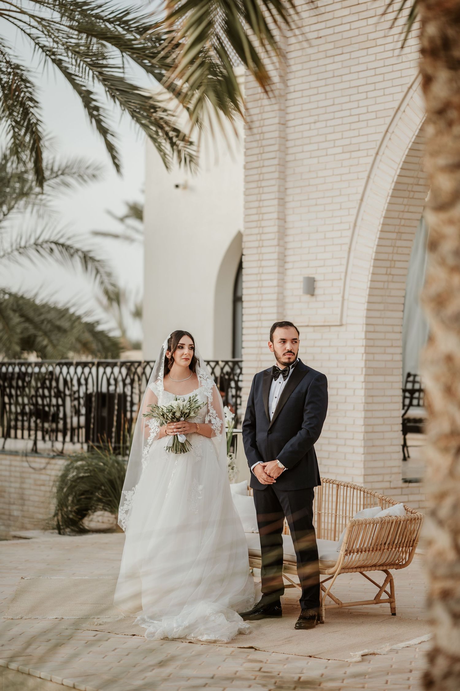 Bride in white gown and groom in tuxedo stand together amid palm trees at Middle Eastern wedding venue.