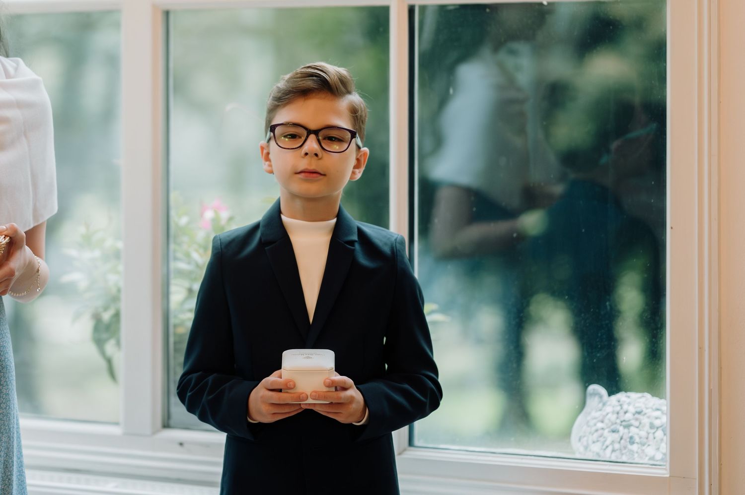 A young boy in a black suit and glasses holds a white candle during a wedding ceremony.