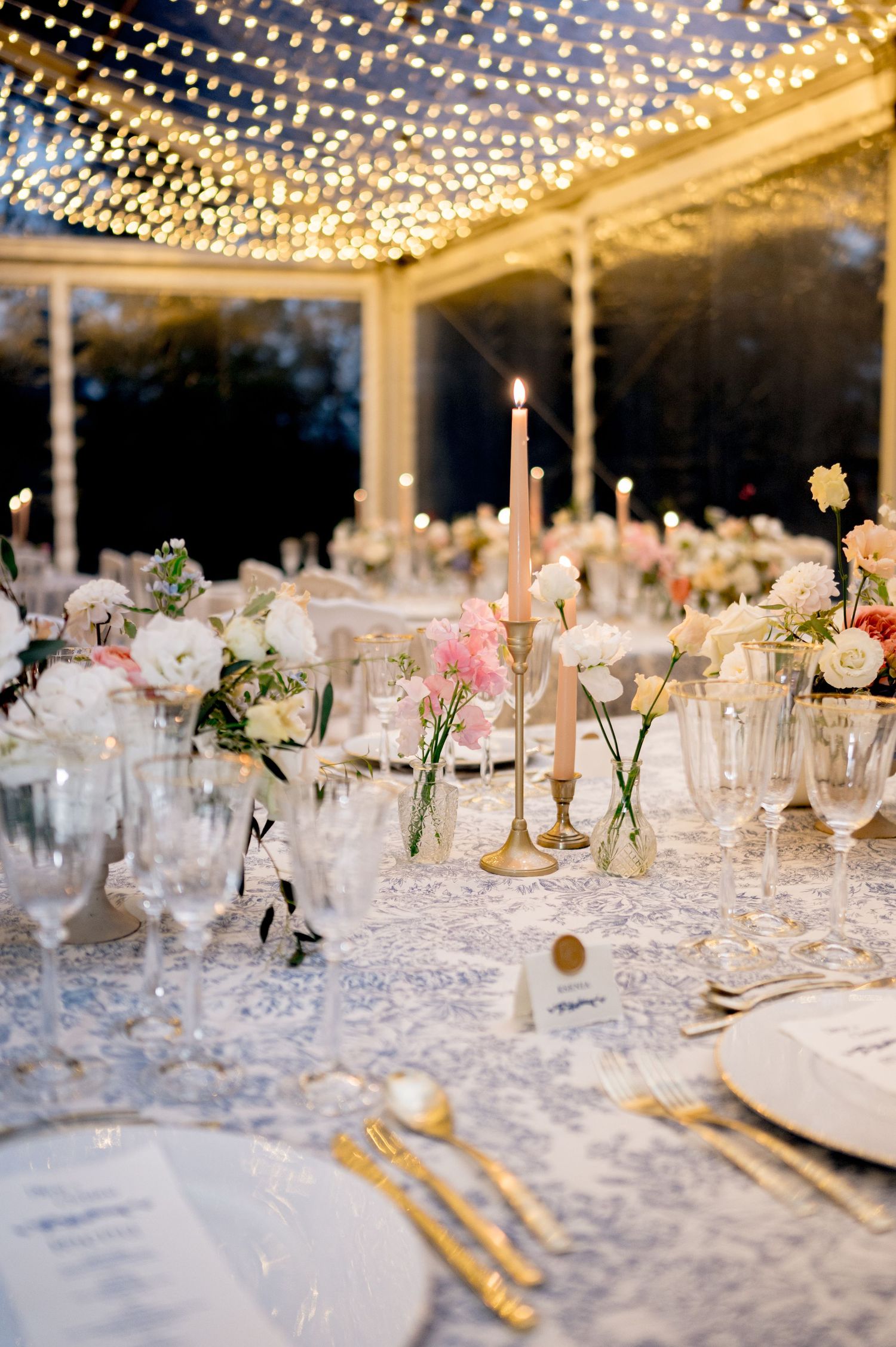 Dreamy wedding dinner table under string lights with pink taper candles, mixed florals, and elegant gold place settings.