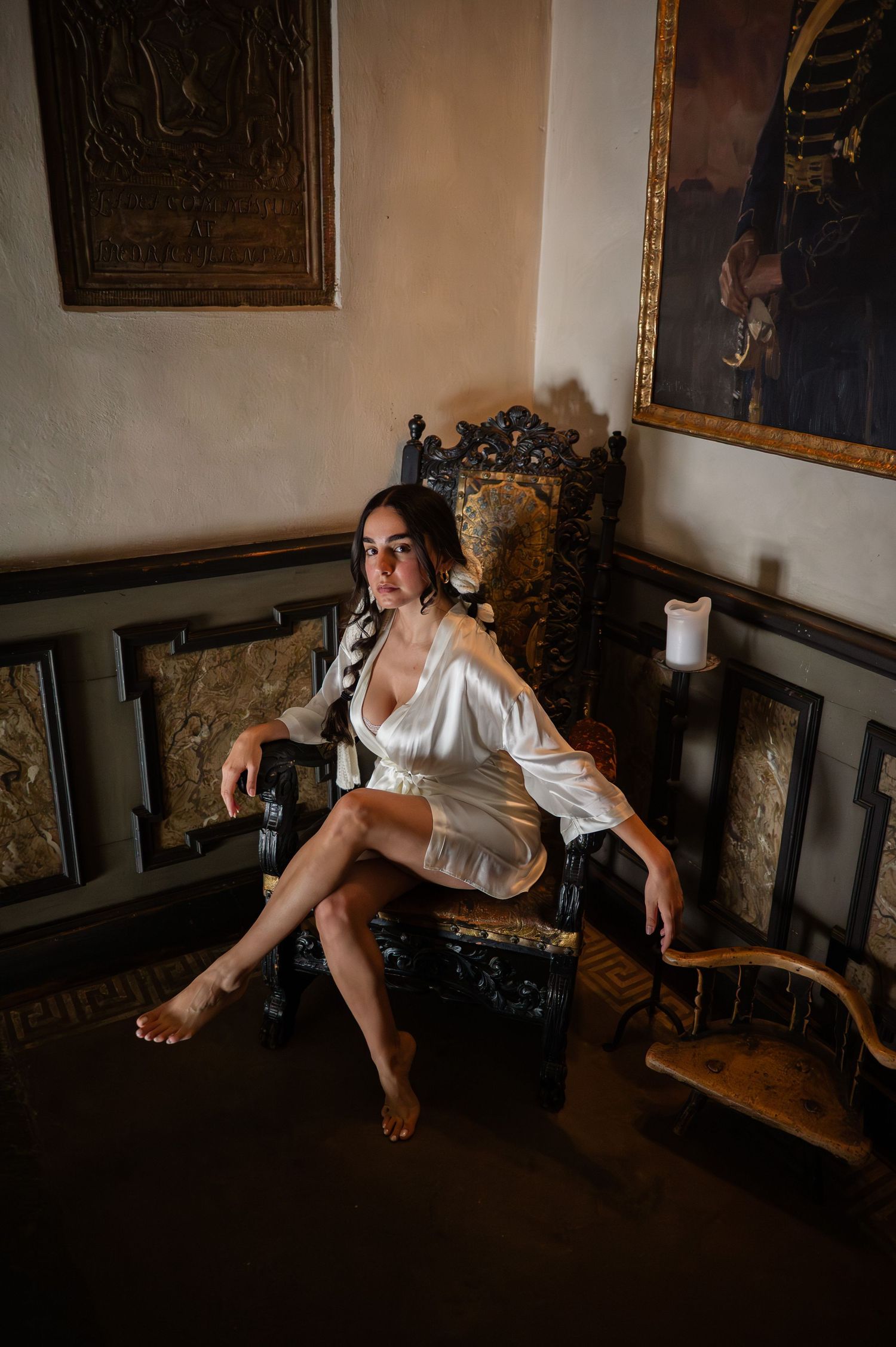 Bride in white silk robe seated on ornate throne chair in dramatic candlelit vintage interior.