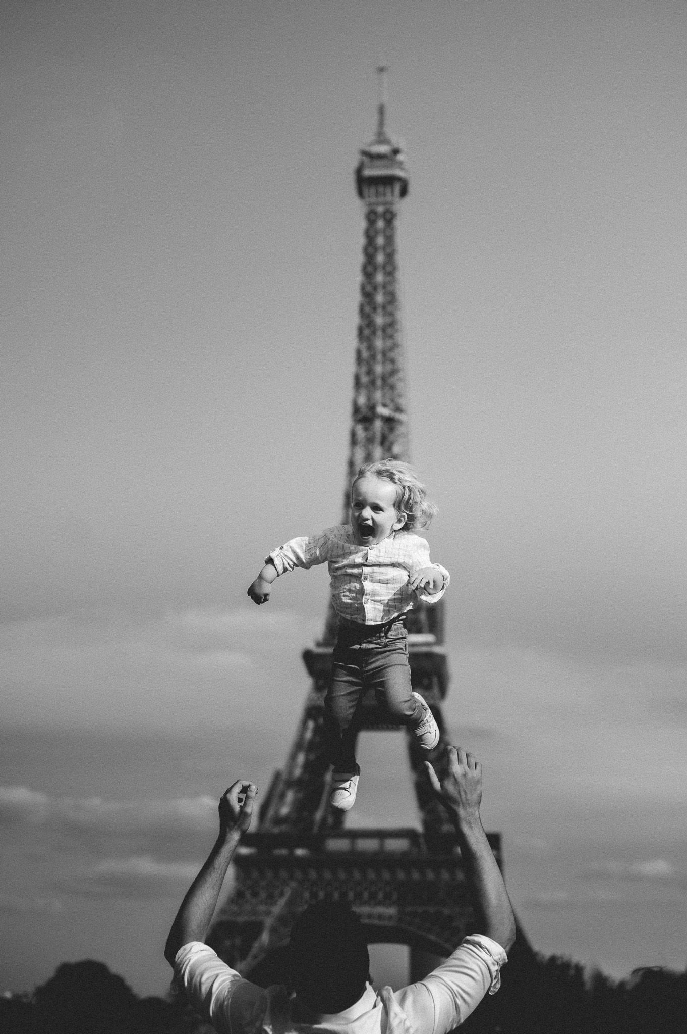 Black and white photo of parent tossing laughing toddler in the air with the Eiffel Tower in background.