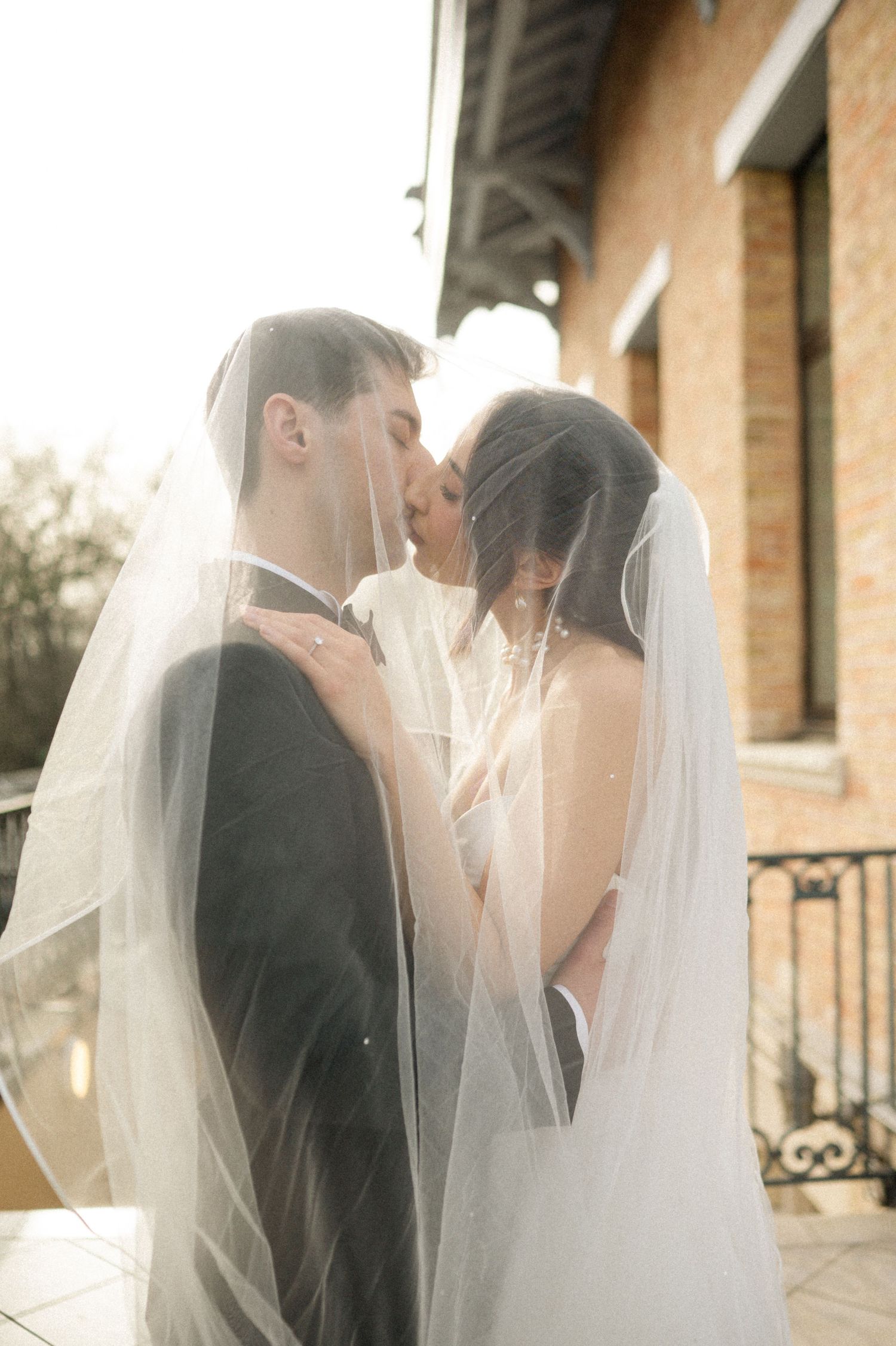 Bride and groom sharing a romantic kiss under a flowing white veil on a sunlit wedding day.