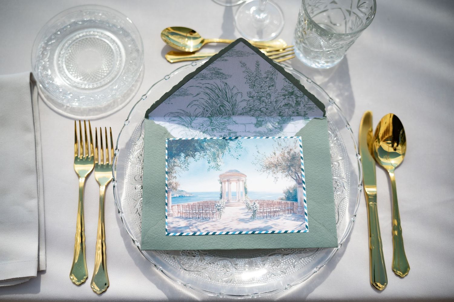 Elegant wedding place setting with gold cutlery, crystal glassware and illustrated venue menu card on white linen.