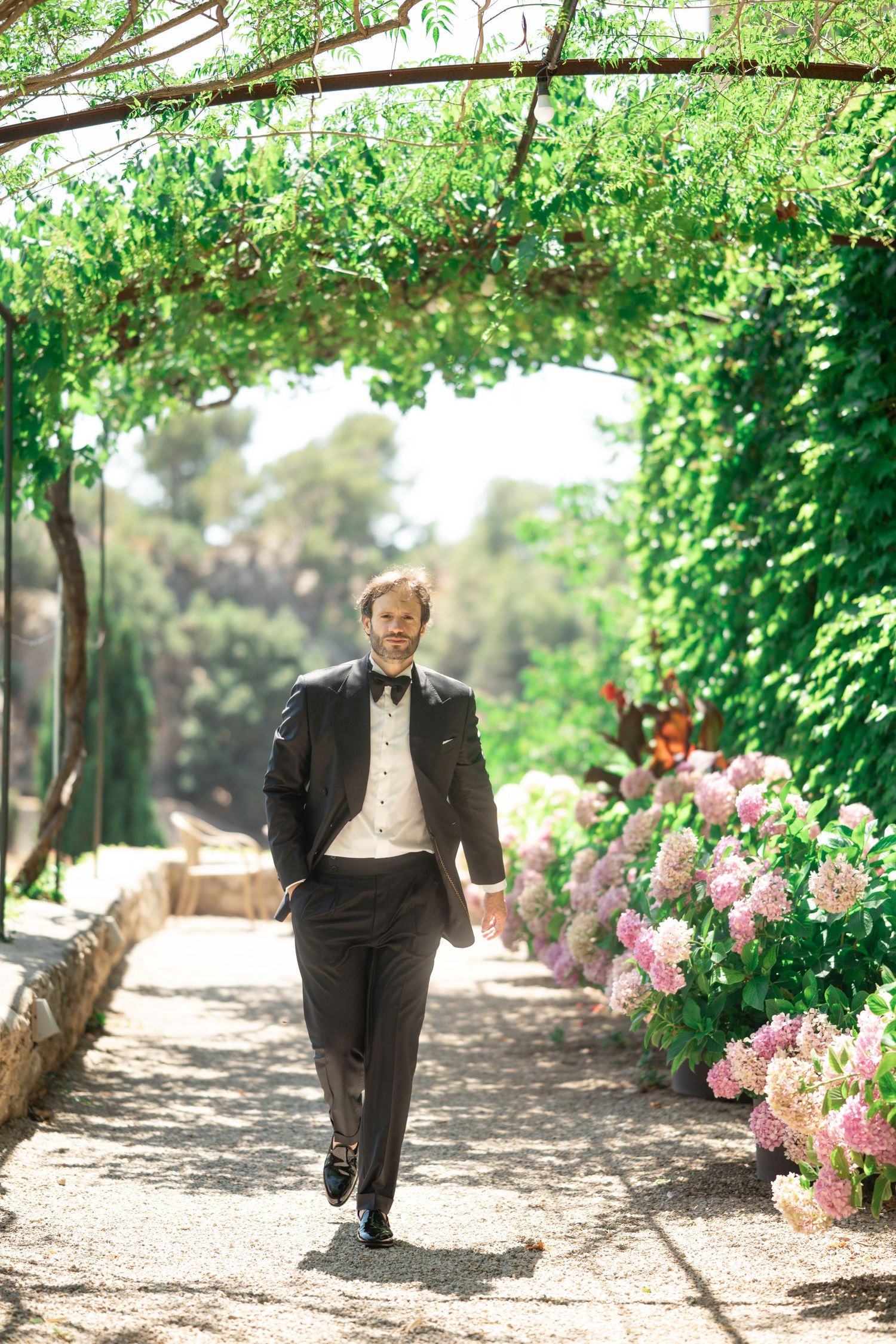 Elegant groom in black tuxedo walks along garden path lined with pink hydrangeas under green archway.
