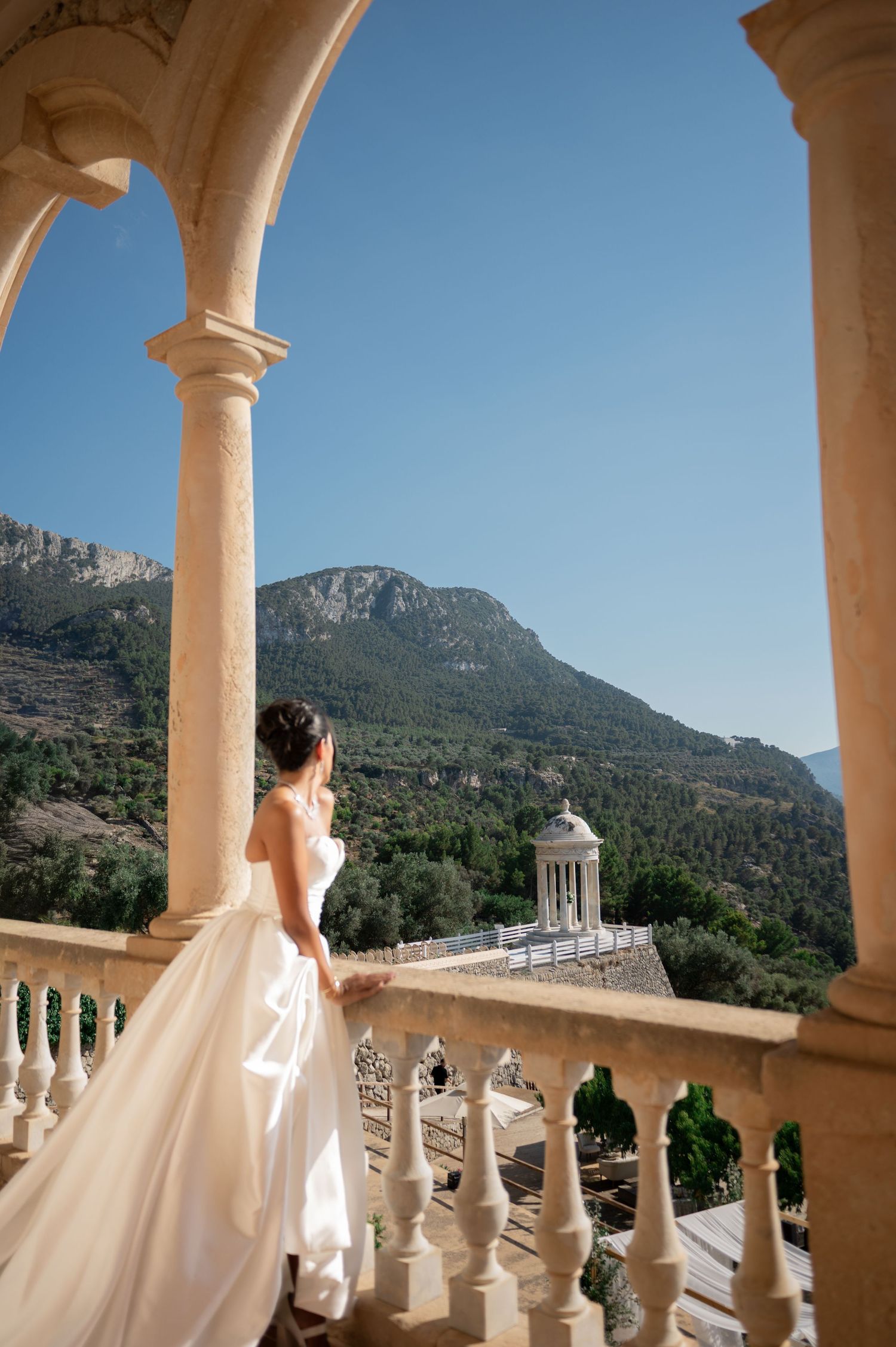 Bride in strapless white gown poses on stone balcony with stunning mountain landscape backdrop.