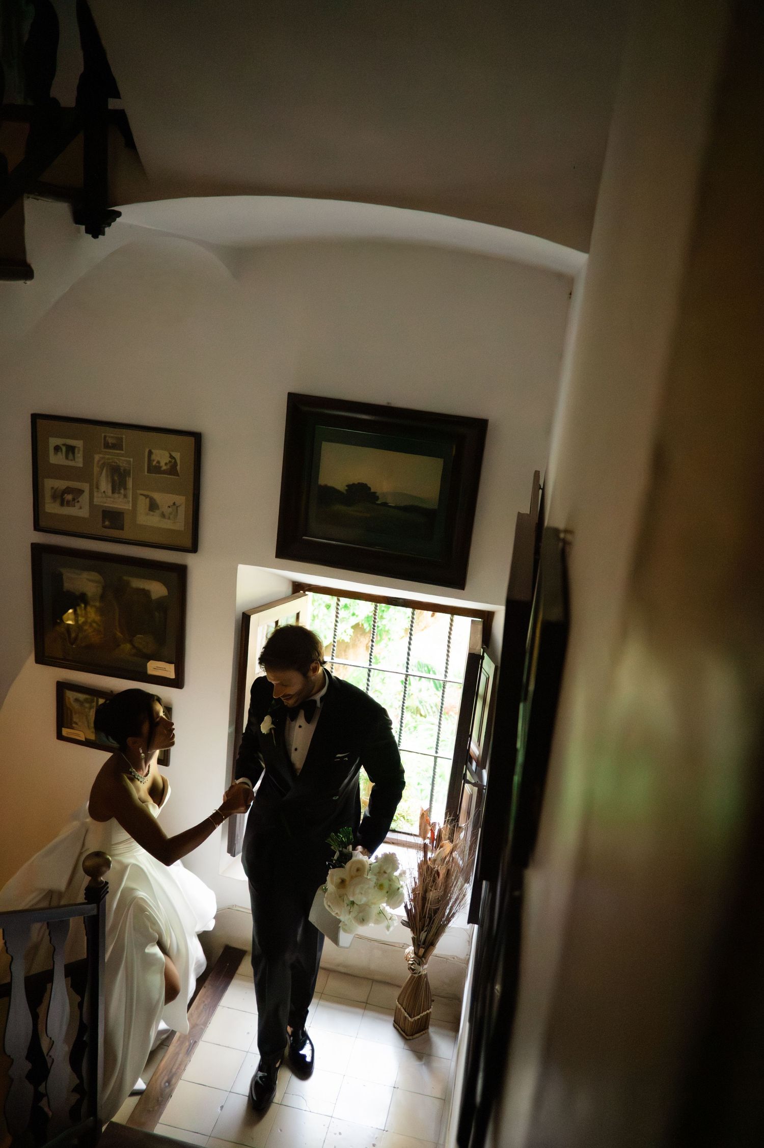 Bride and groom ascend staircase inside historic villa adorned with framed artwork on walls.