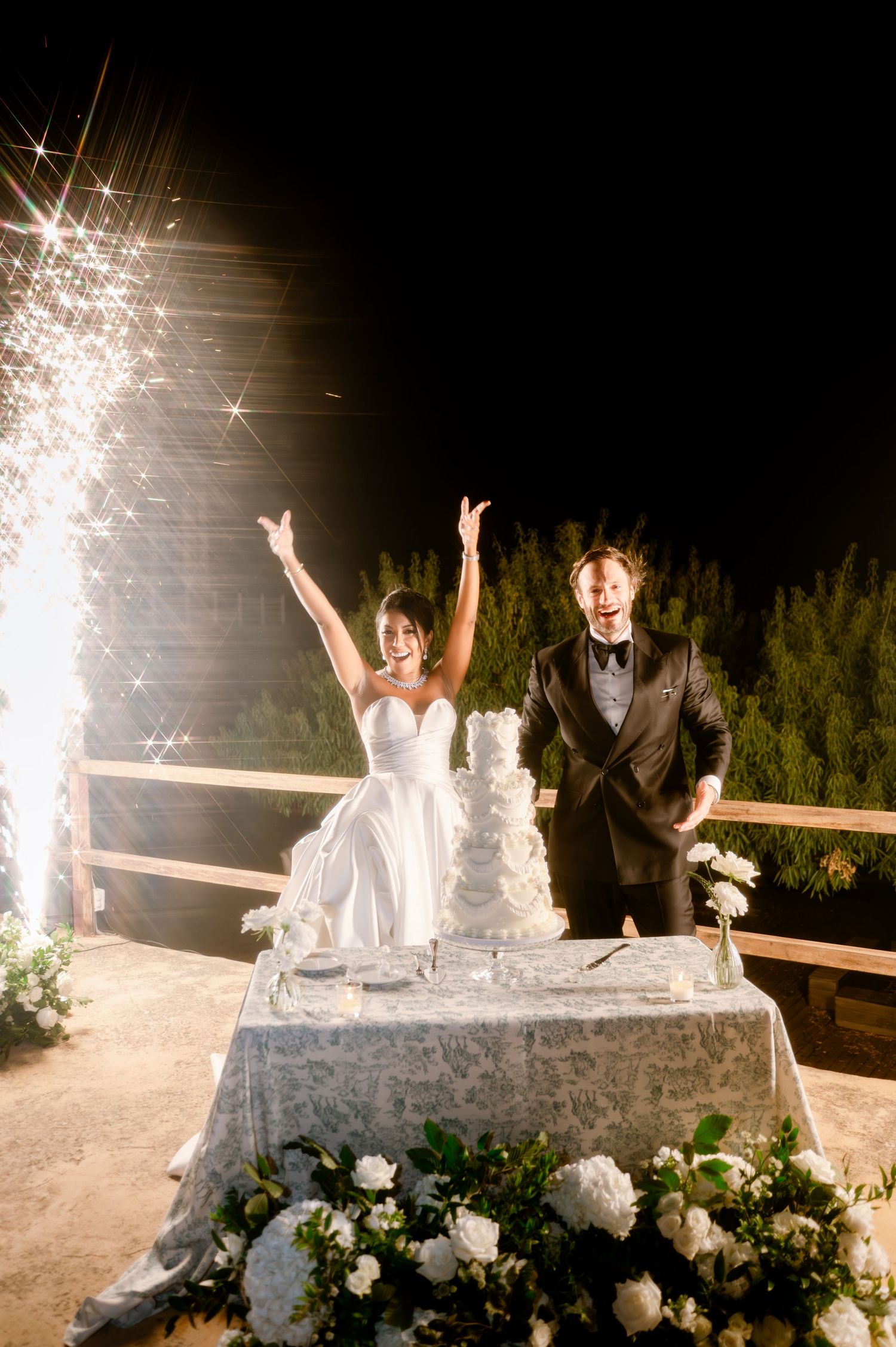 Excited bride and groom raise hands in celebration beside wedding cake with firework sparklers at night.