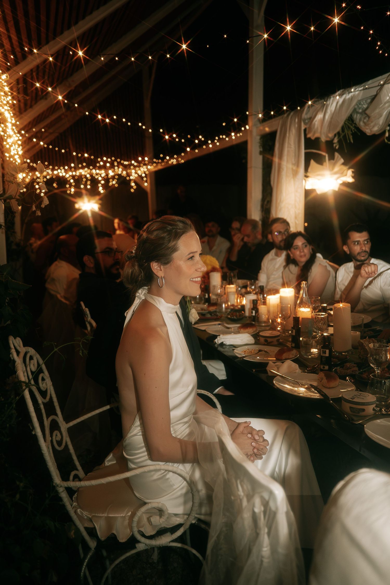 Radiant bride in white satin gown seated at candlelit outdoor wedding reception under string lights.