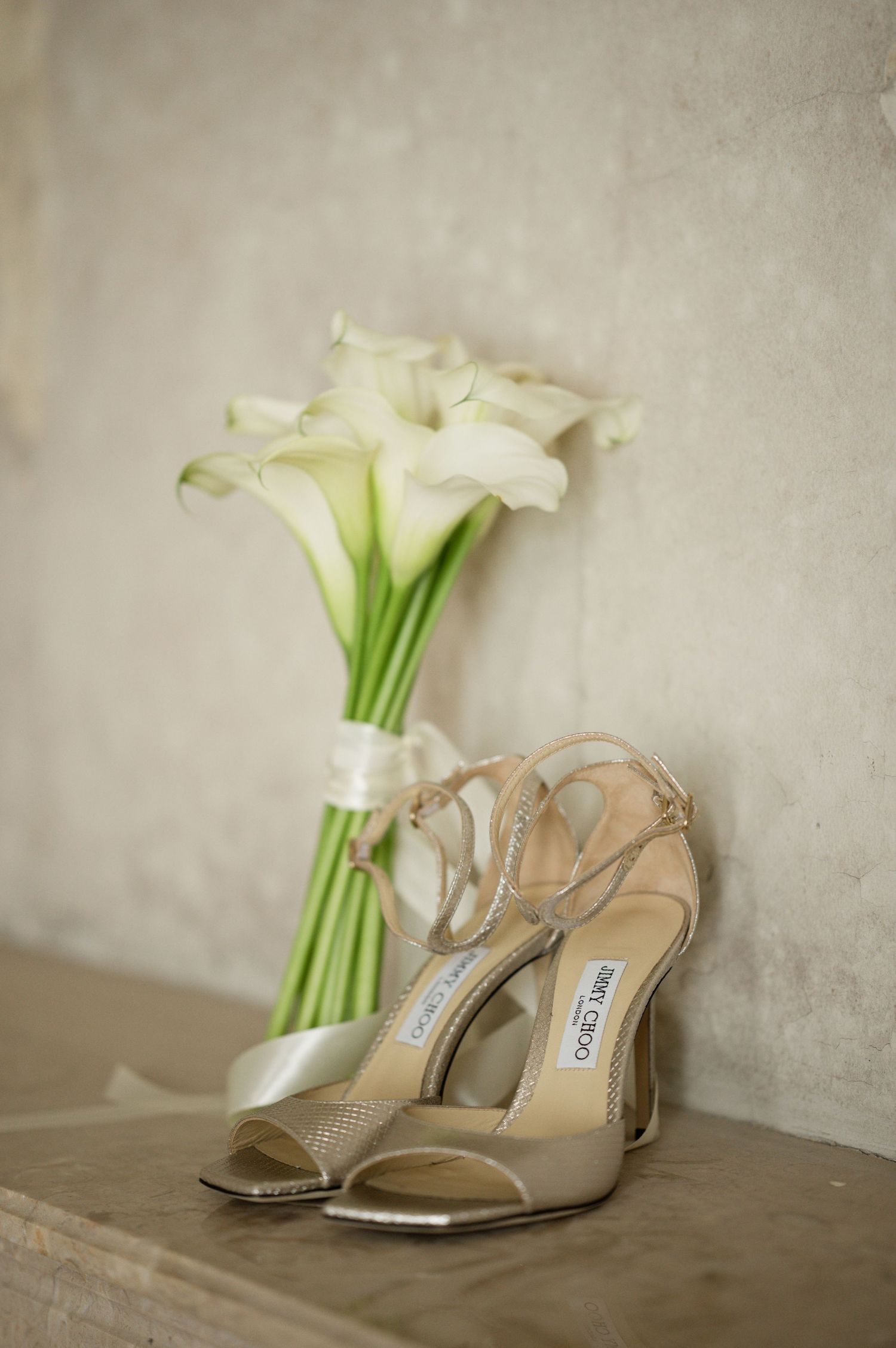 Jimmy Choo bridal heels beside white calla lily bouquet on stone surface.