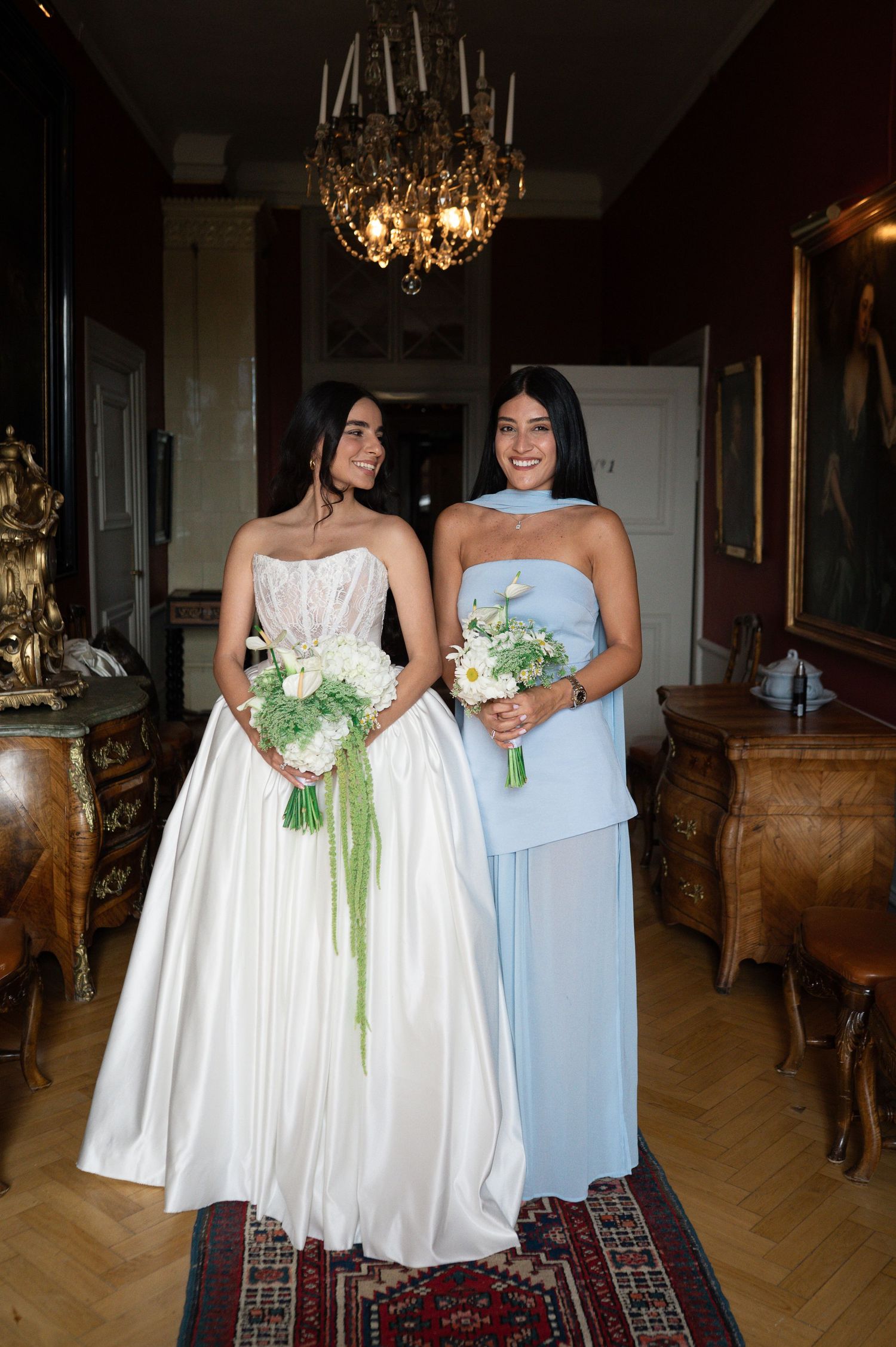 Bride in strapless white ball gown and bridesmaid in blue dress holding bouquets in ornate room.