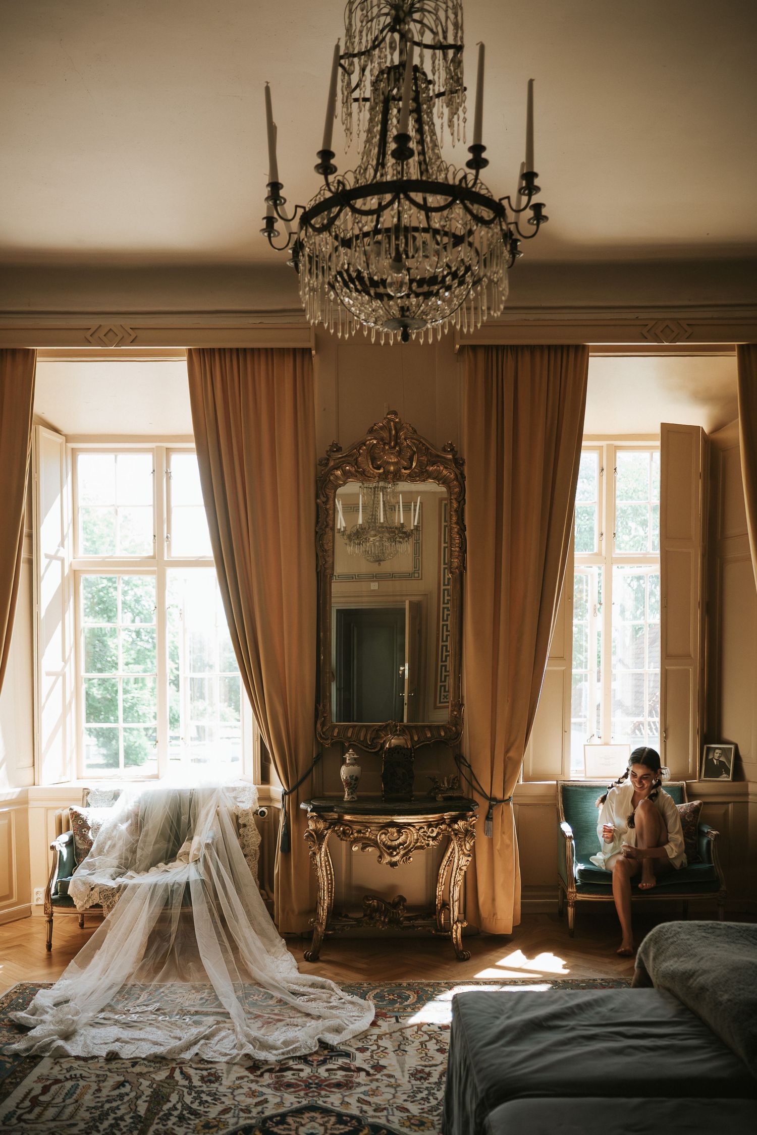 Elegant vintage bridal suite with crystal chandelier, ornate mirror, and flowing wedding veil draped over furniture.