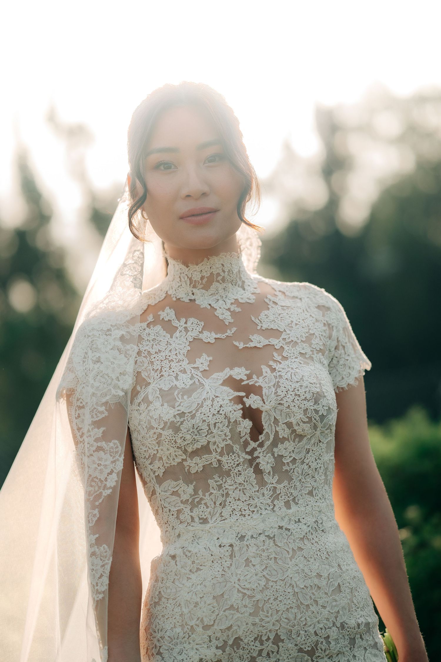 Beautiful bride in high-neck lace wedding gown with veil in golden backlit sunlight.
