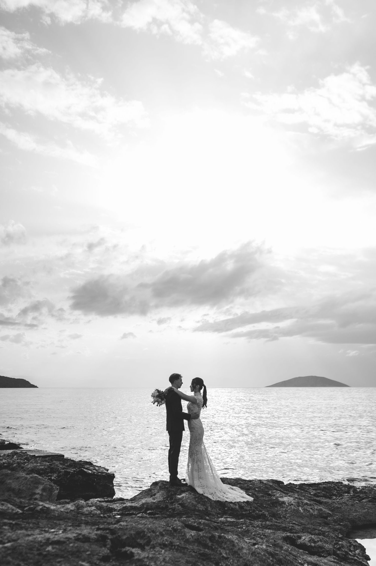 Bride and groom share a romantic silhouette moment on rocky coast with dramatic sunlit sky and sea.