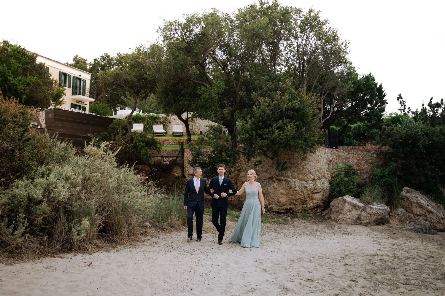 Three wedding guests walk along sandy beach path surrounded by lush greenery and stone wall.