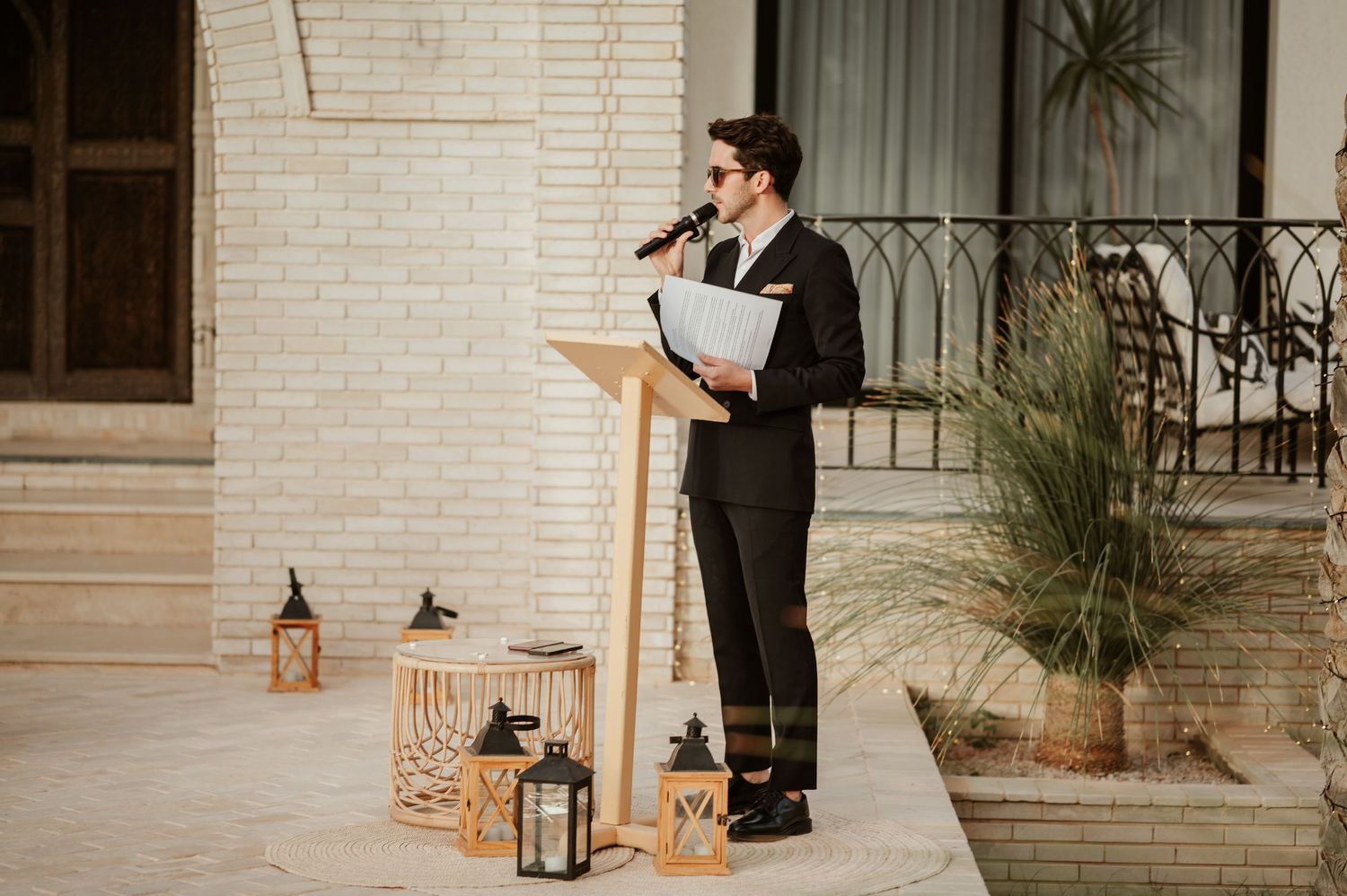 Dapper officiant in black suit reads from papers at a wooden podium flanked by lanterns at outdoor wedding.