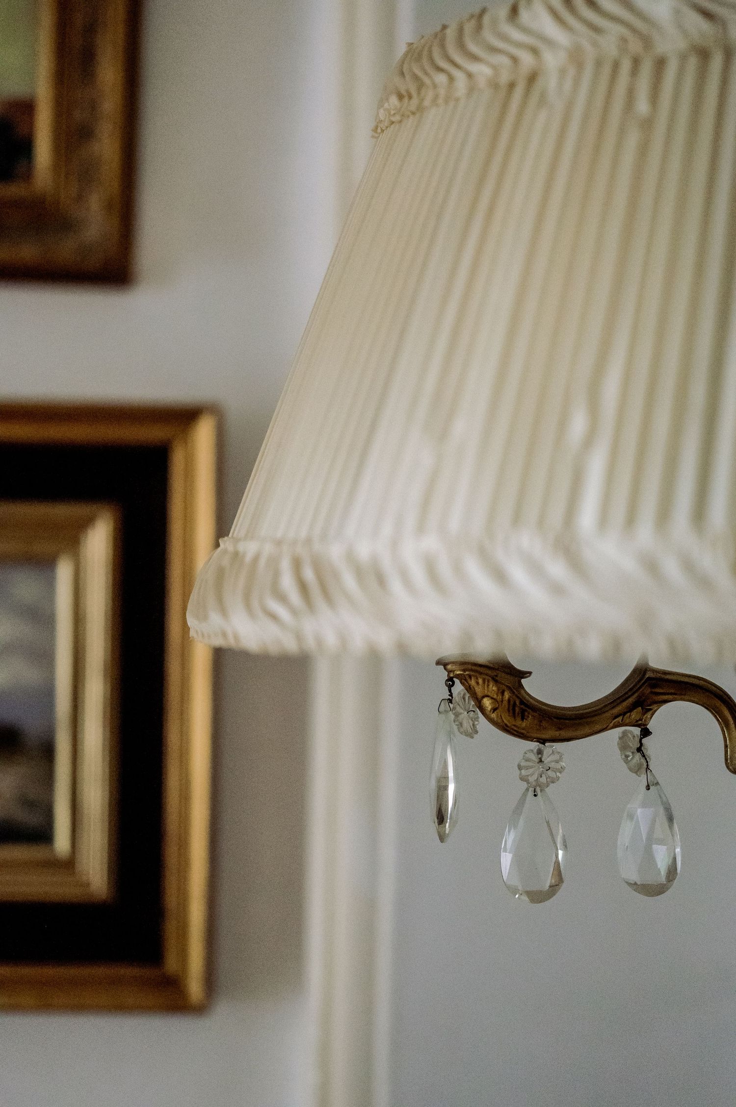 Close-up of an antique brass lamp with pleated cream shade and crystal drop pendants beside gold-framed paintings.