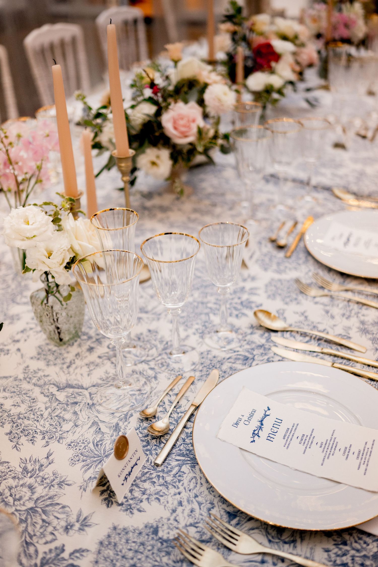 Luxury wedding place setting with gold cutlery, crystal glasses, blue toile linen, and menu card on white charger plate.