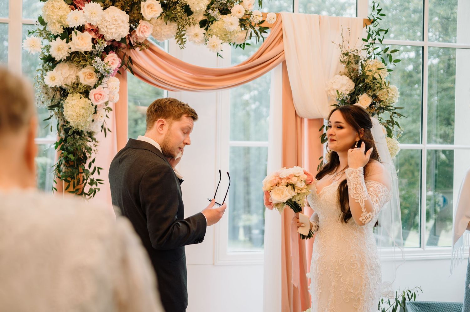 Groom reads vows to emotional bride beneath elegant pink and white floral wedding arch.