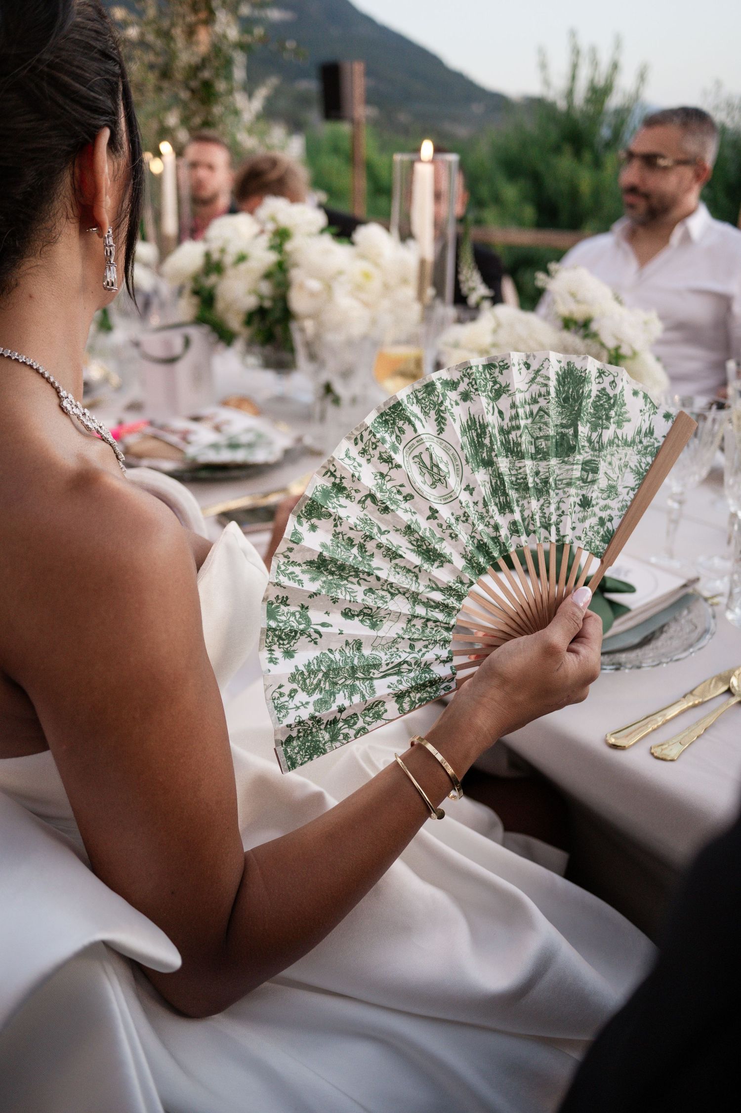 Close-up of bride holding ornate green floral hand fan at elegant outdoor wedding reception dinner.