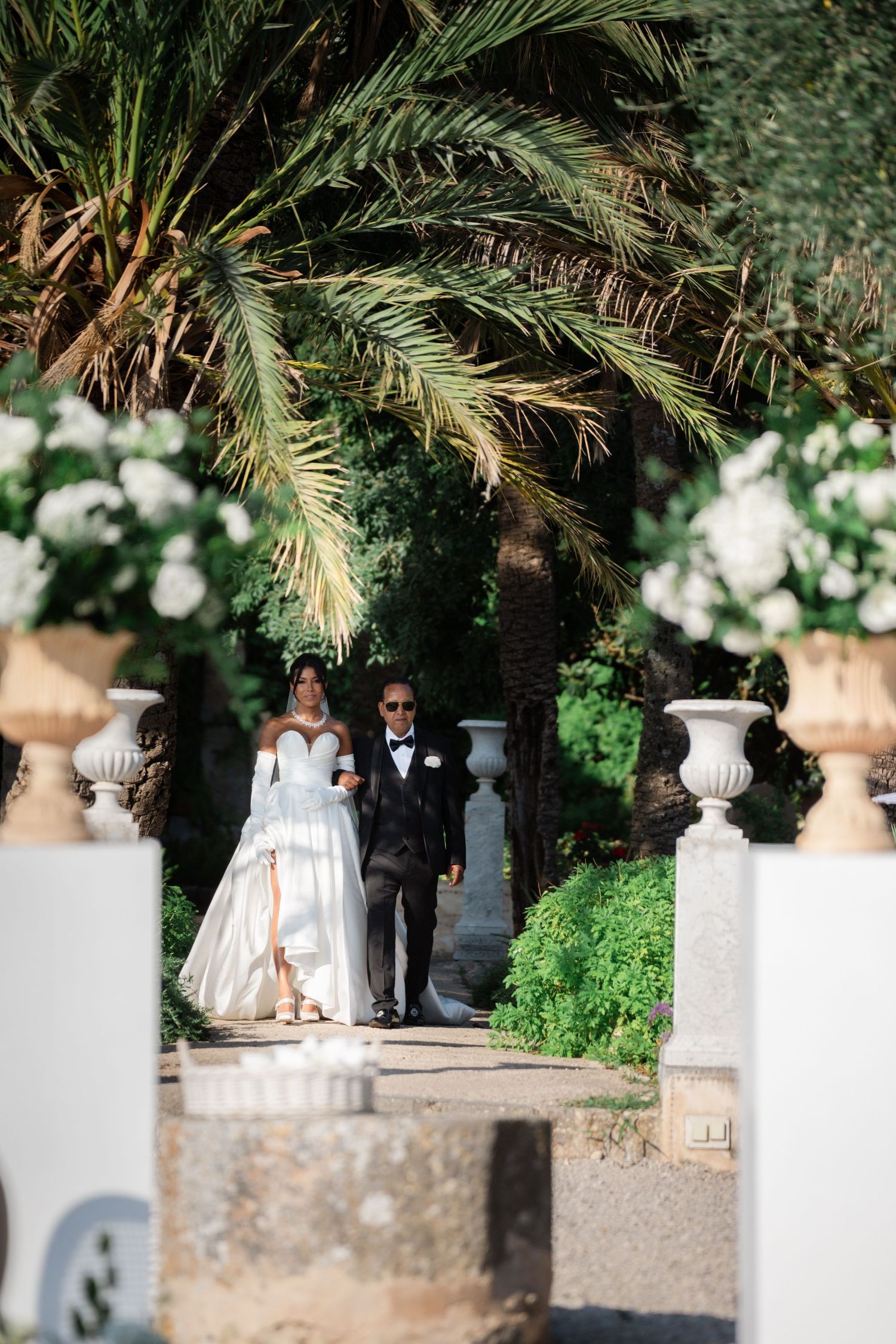 Bride in a white off-shoulder gown walks down the aisle with her father through a garden path.