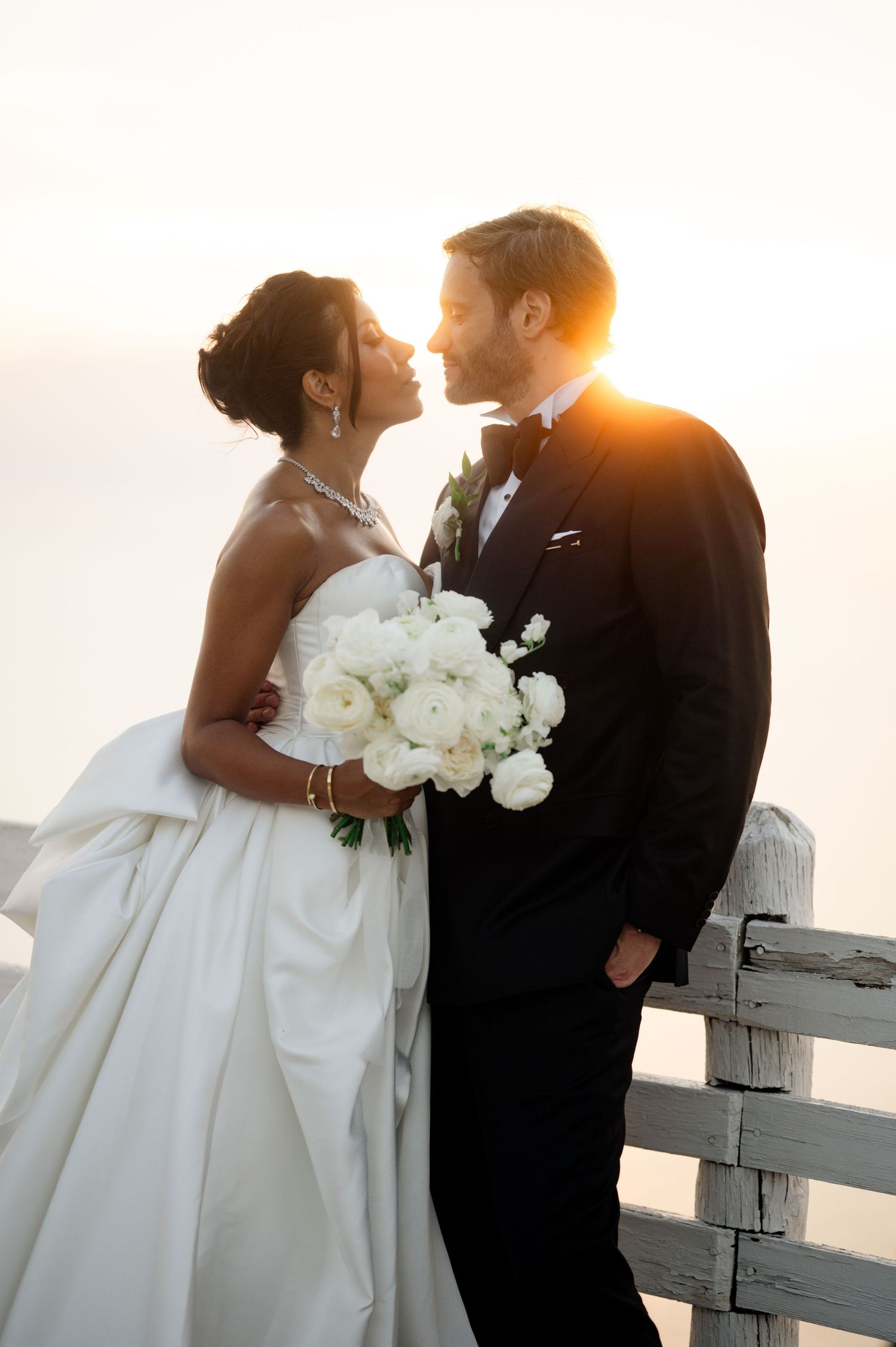 Bride and groom share intimate moment holding white peony bouquet with stunning golden sunset backdrop.
