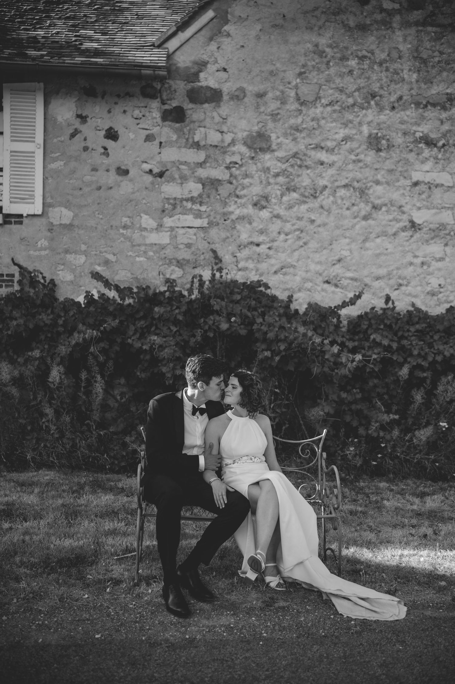 Black and white romantic wedding photo of bride and groom embracing on decorative bench against ivy-covered stone wall.
