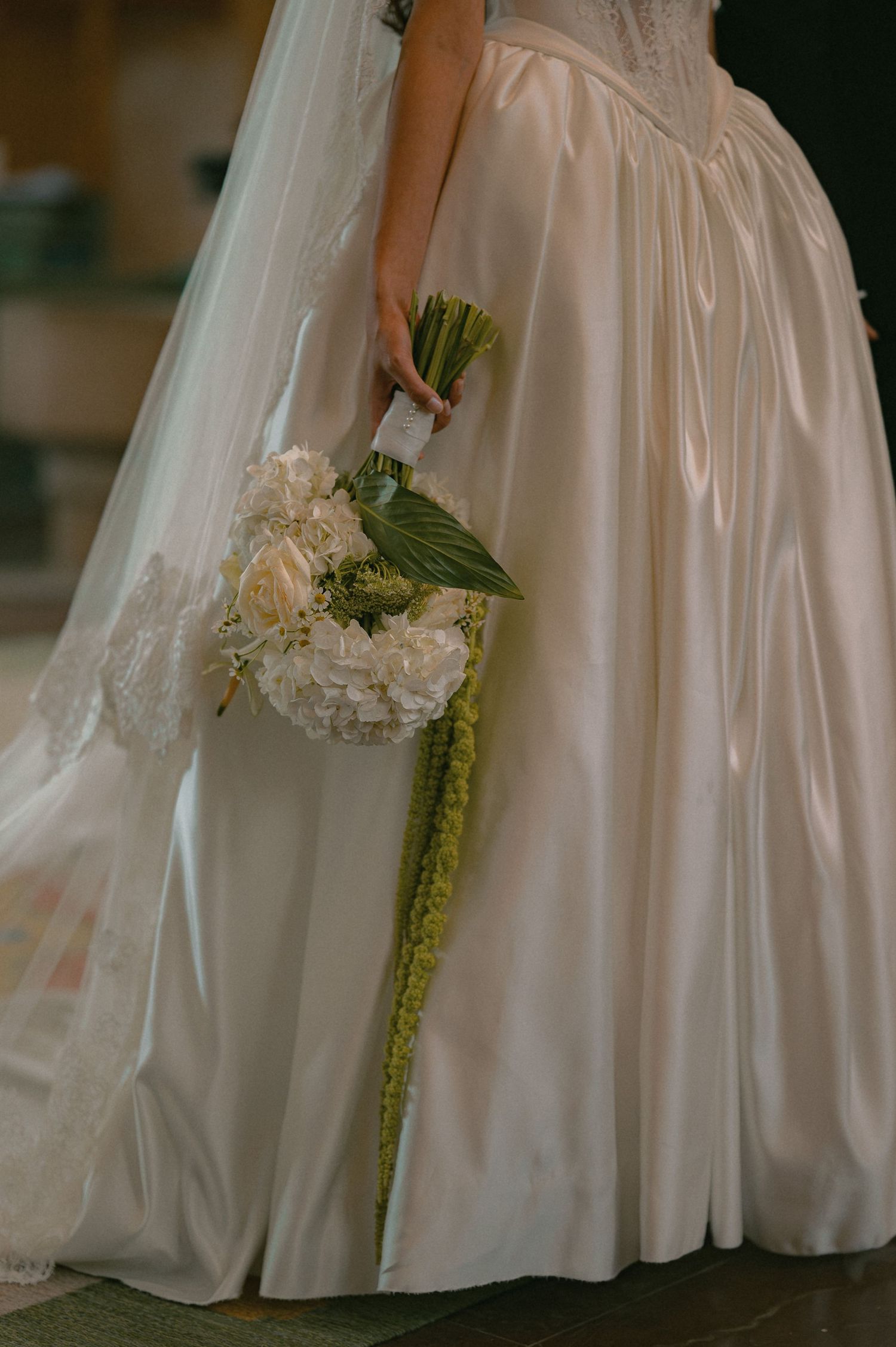 Bride in ivory satin ballgown holds white hydrangea bouquet with trailing green amaranthus stems.