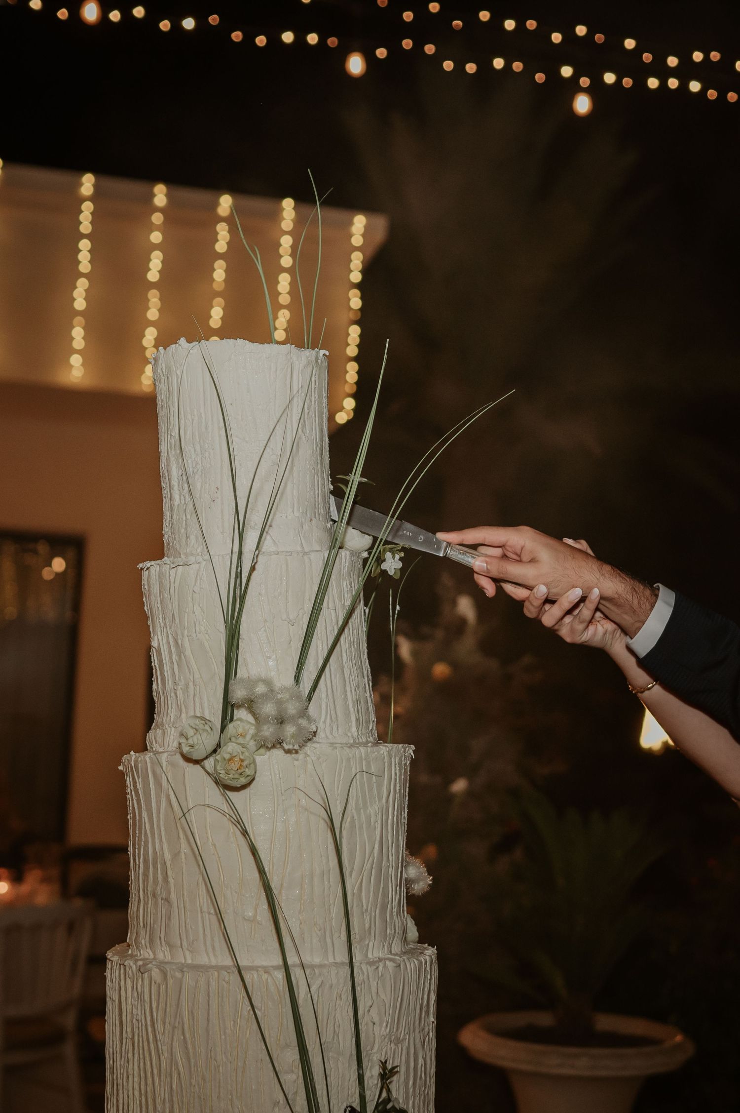 Close-up of hands cutting elegant four-tier textured white wedding cake decorated with green stems outdoors.