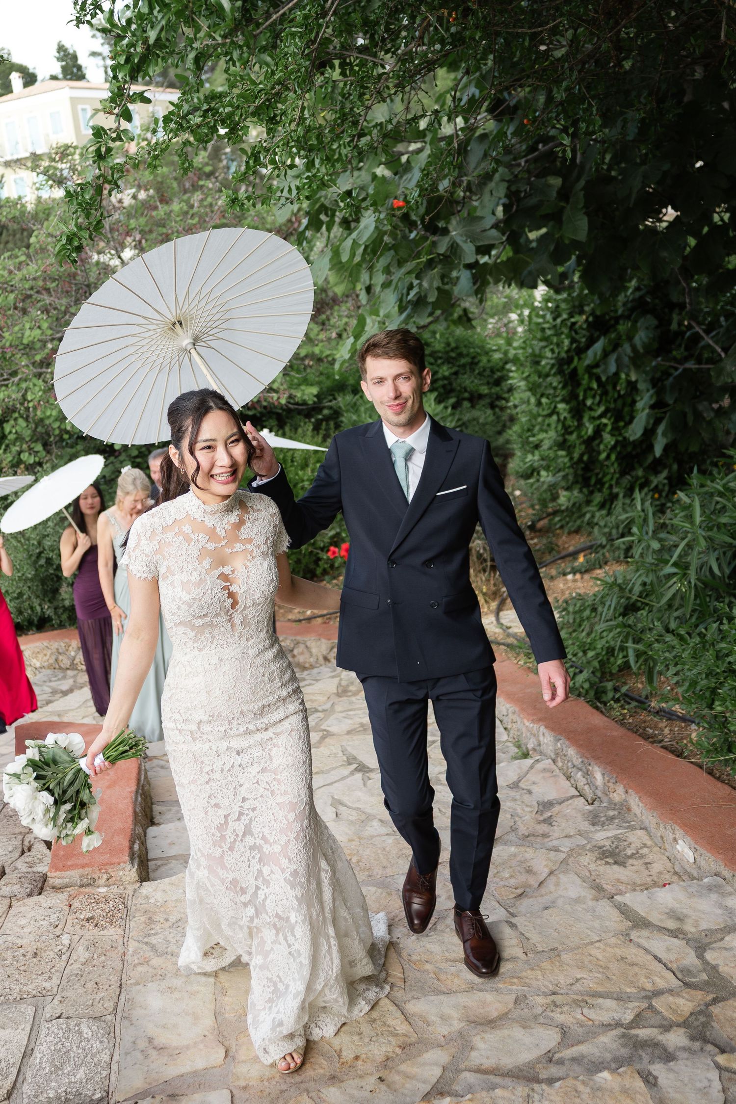 Smiling newlyweds walk under white parasols through lush garden after wedding ceremony.