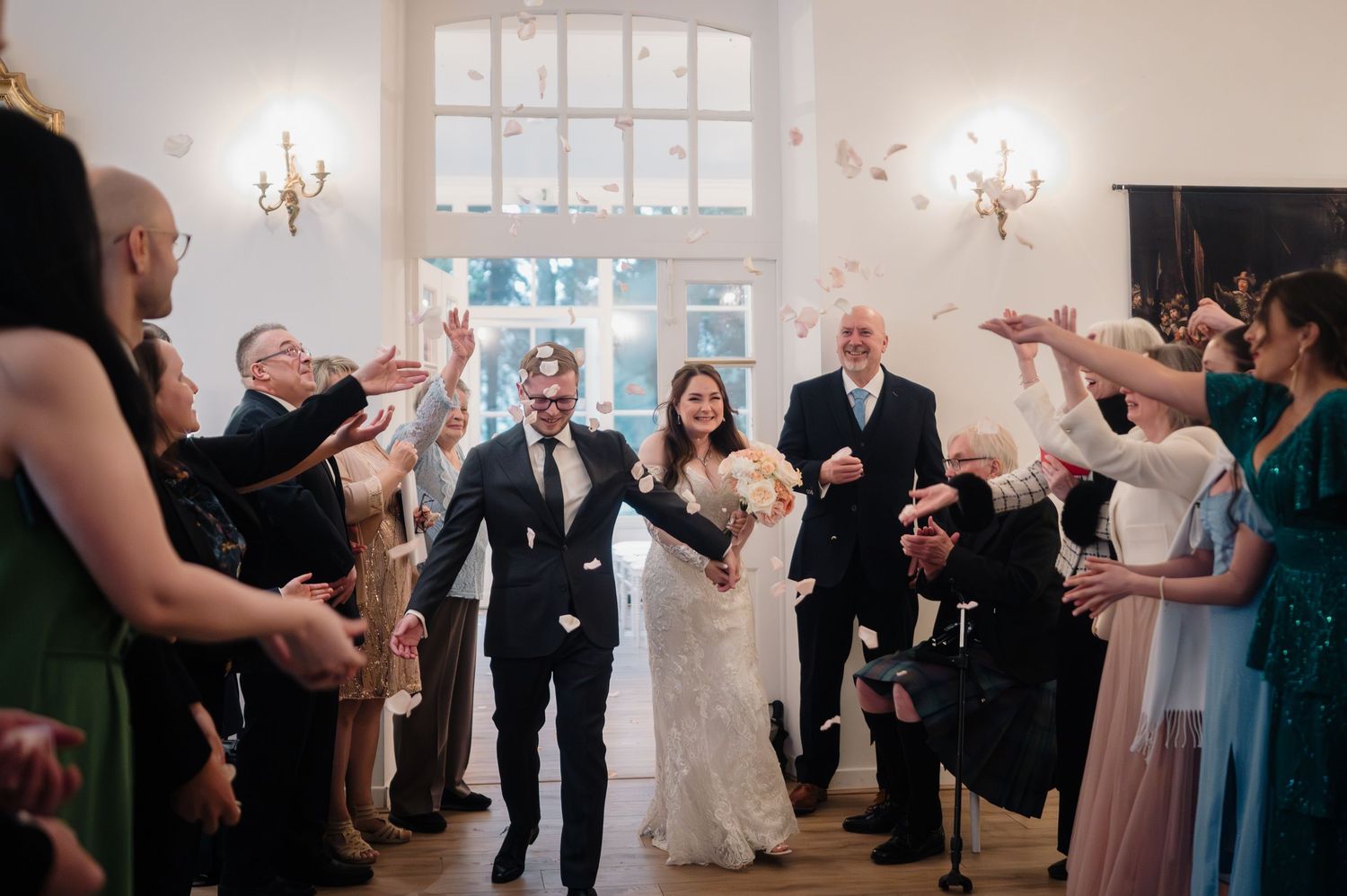 Bride and groom walk through cheering guests throwing confetti in elegant white wedding venue.