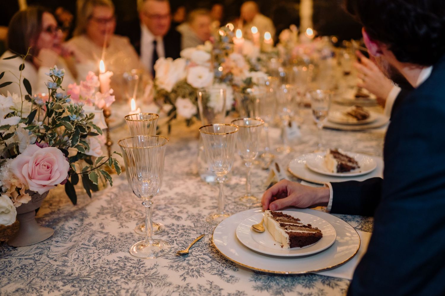 Elegant wedding reception dinner table with guests enjoying cake beside crystal champagne flutes and flowers.