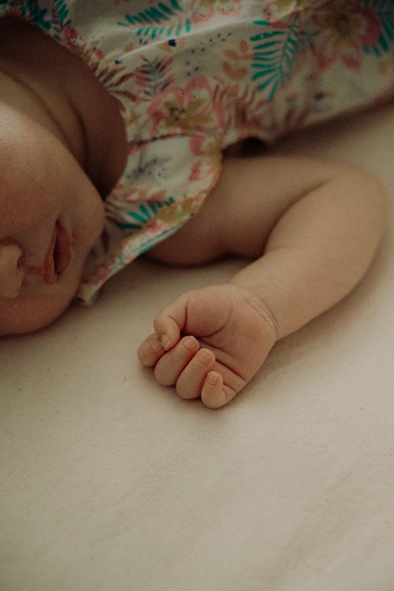 Sleeping newborn baby in floral outfit with tiny fist resting on white surface in soft natural light.