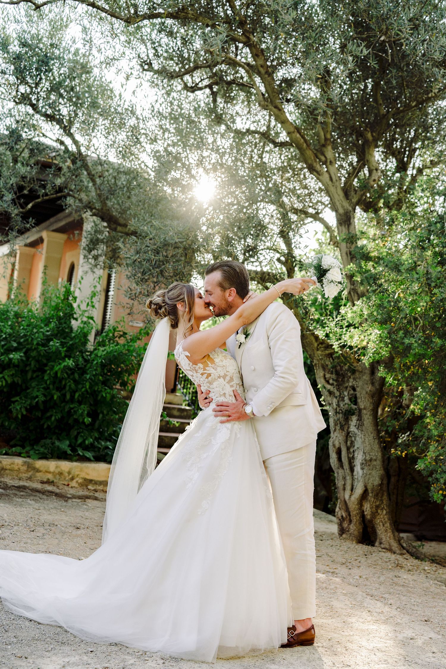 Bride and groom embrace and kiss beneath sunlit olive trees in a romantic garden setting.