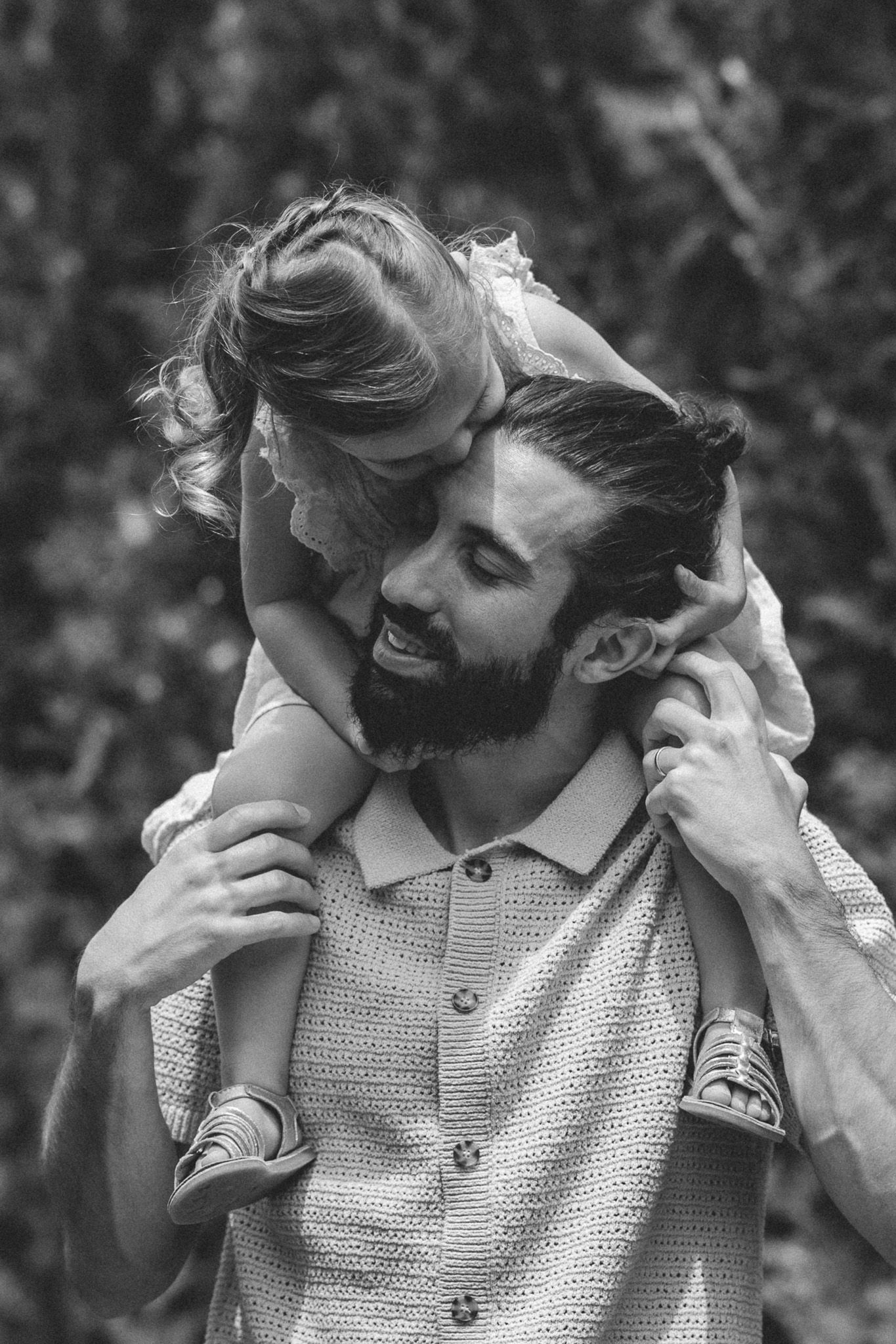 Black and white photo of bearded father smiling with young daughter on his shoulders.