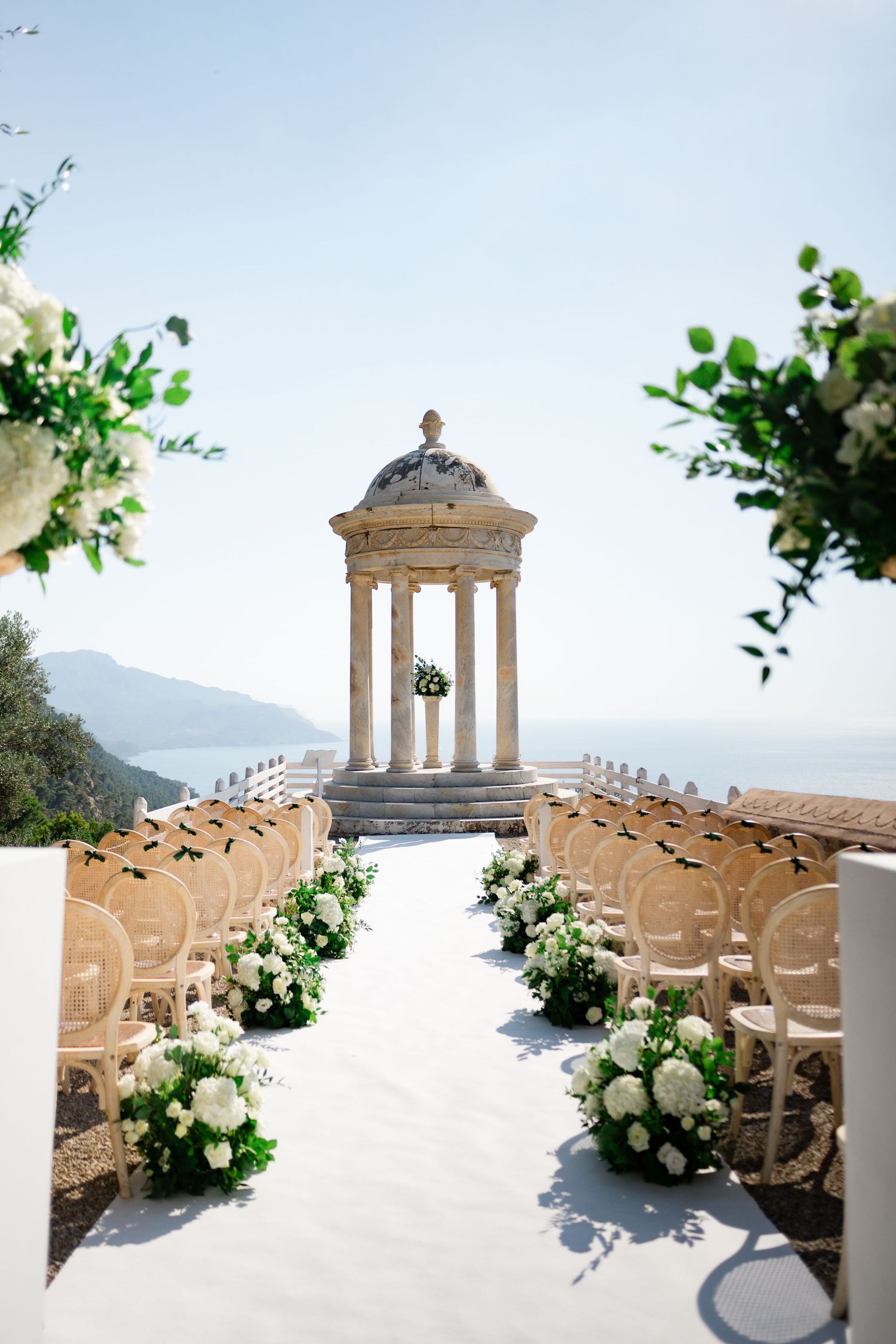 Elegant outdoor wedding ceremony aisle with white floral arrangements leading to a classical stone gazebo overlooking the sea.