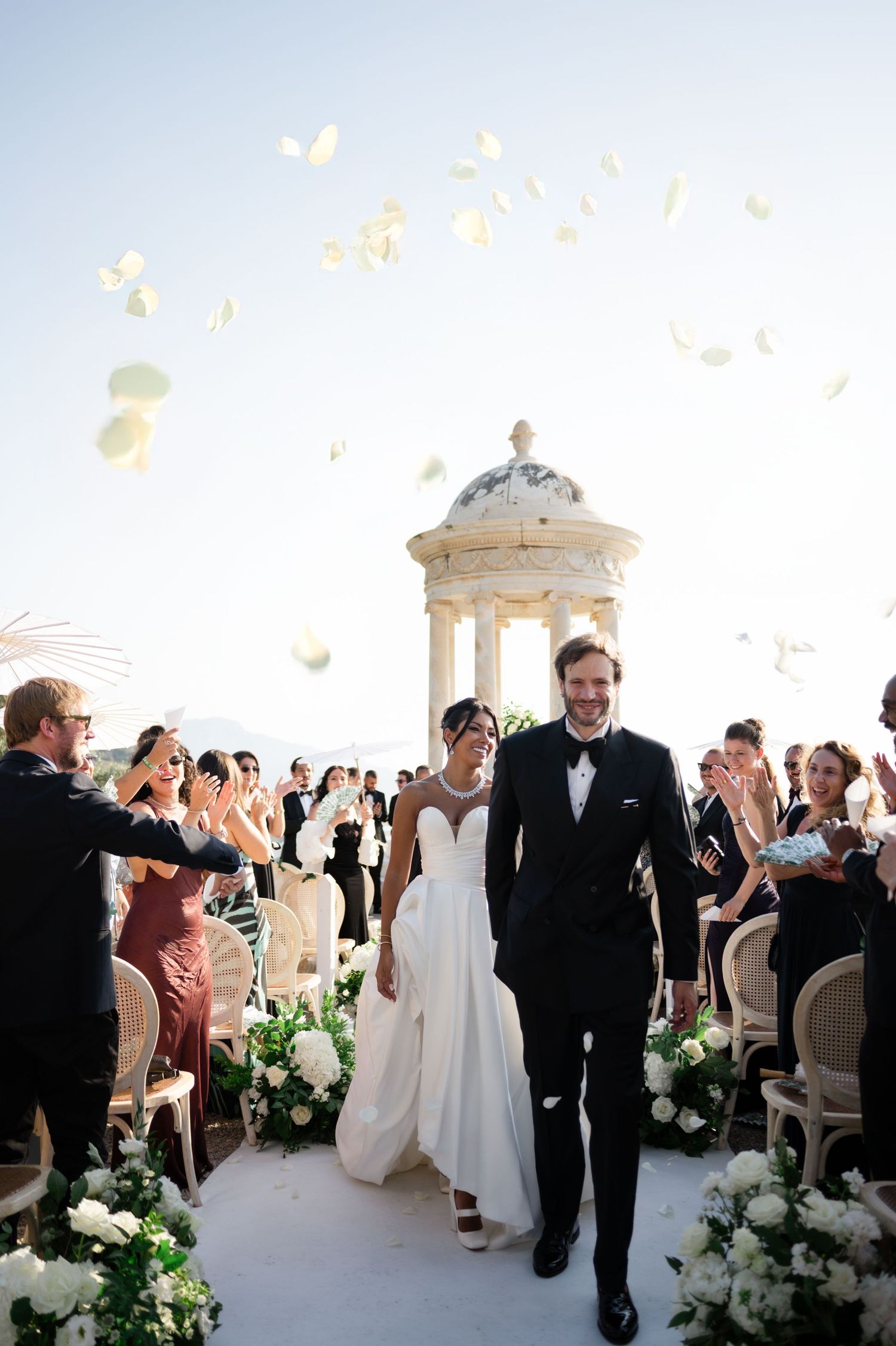 Joyful newlyweds exit ceremony as guests toss white flower petals at stunning outdoor wedding.