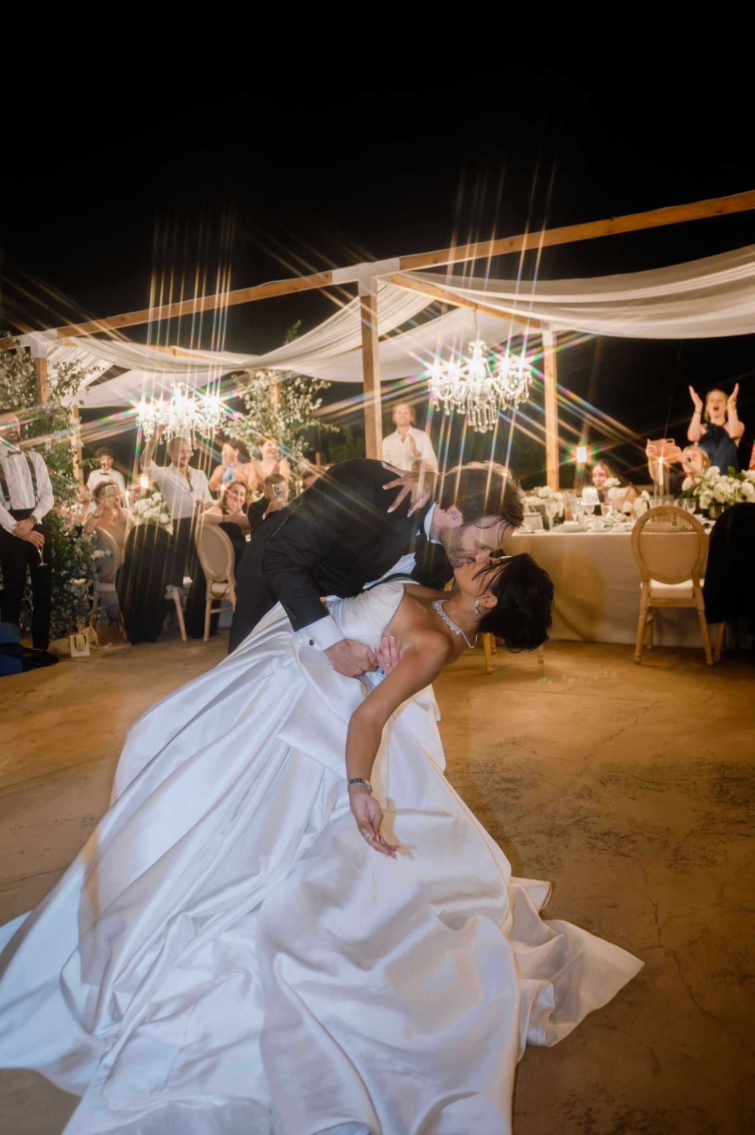Dramatic wedding first dance dip as groom bends bride back for a kiss under chandelier-lit reception tent.