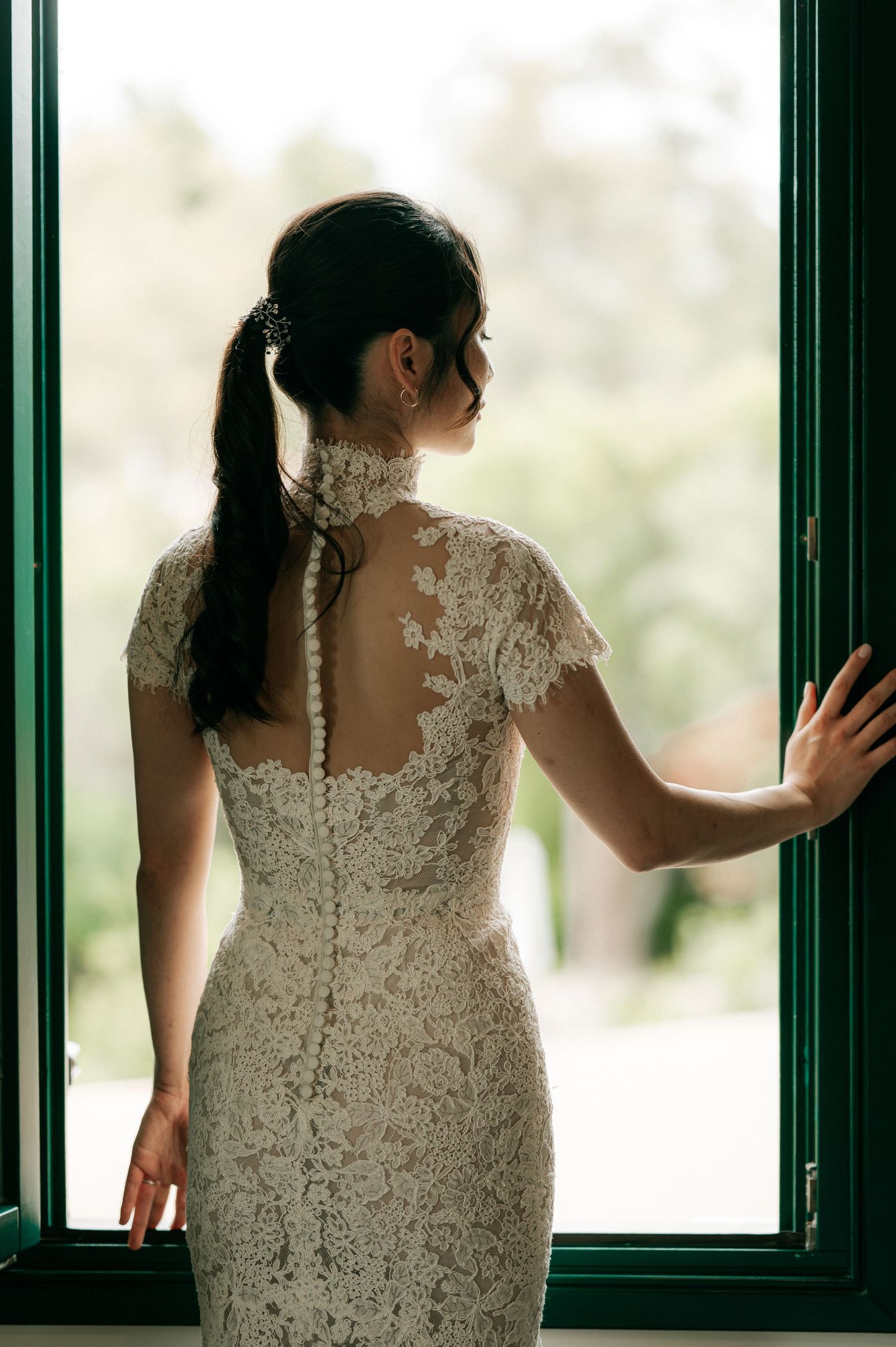 Bride in fitted lace wedding dress with high neck and button back detail standing at an open window.