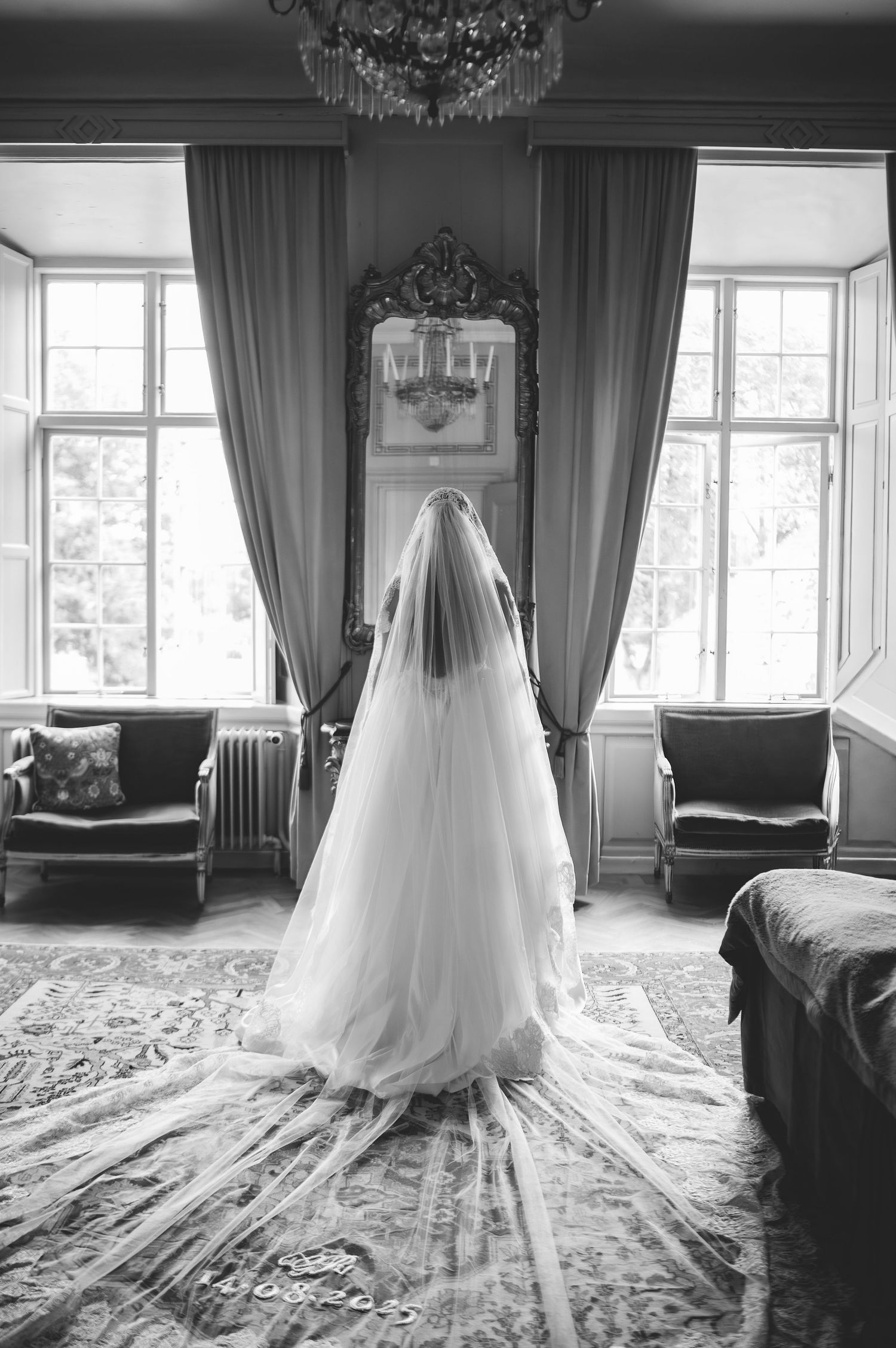 Black and white back view of bride in cathedral veil standing between tall windows in vintage room.