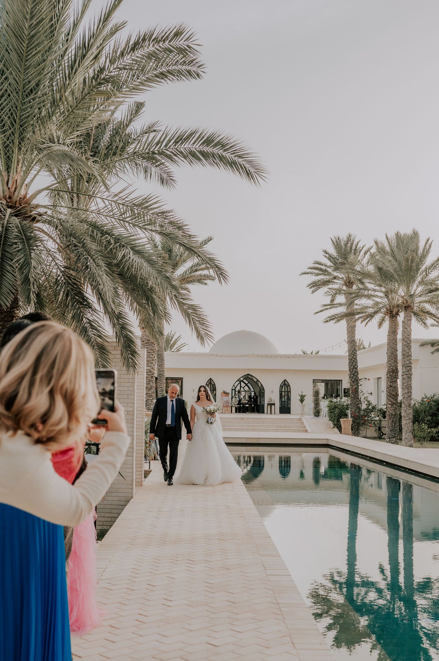 Bride and father walking along a palm-lined reflecting pool as a guest photographs the processional moment.