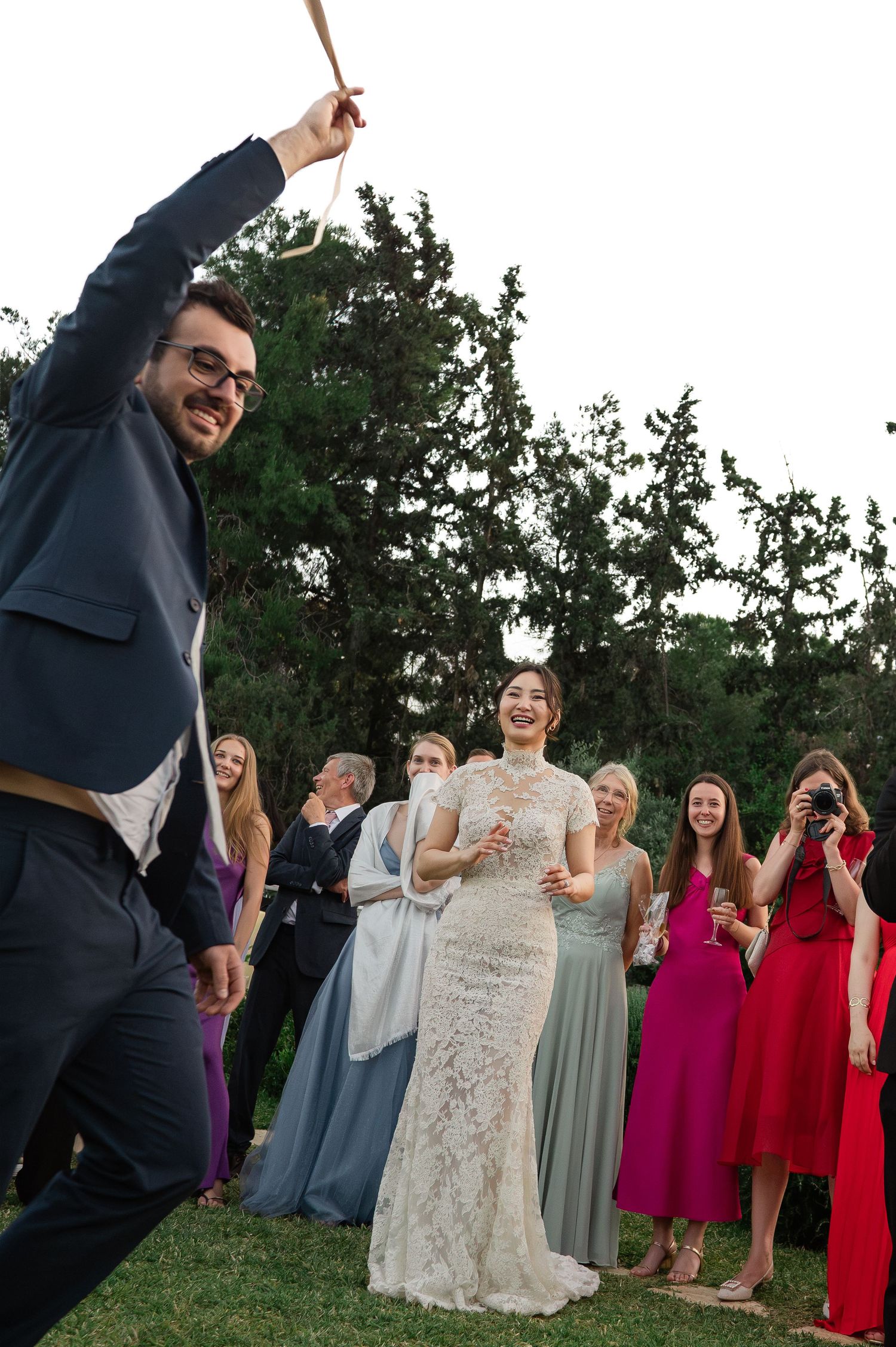 Joyful bride in lace gown celebrates with guests outdoors as groom raises a sparkler at wedding reception.
