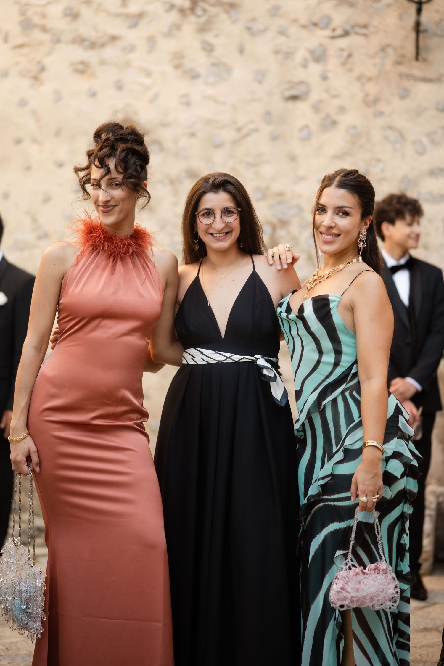 Three women pose together in elegant gowns at a wedding, wearing rose, black, and zebra-print dresses.