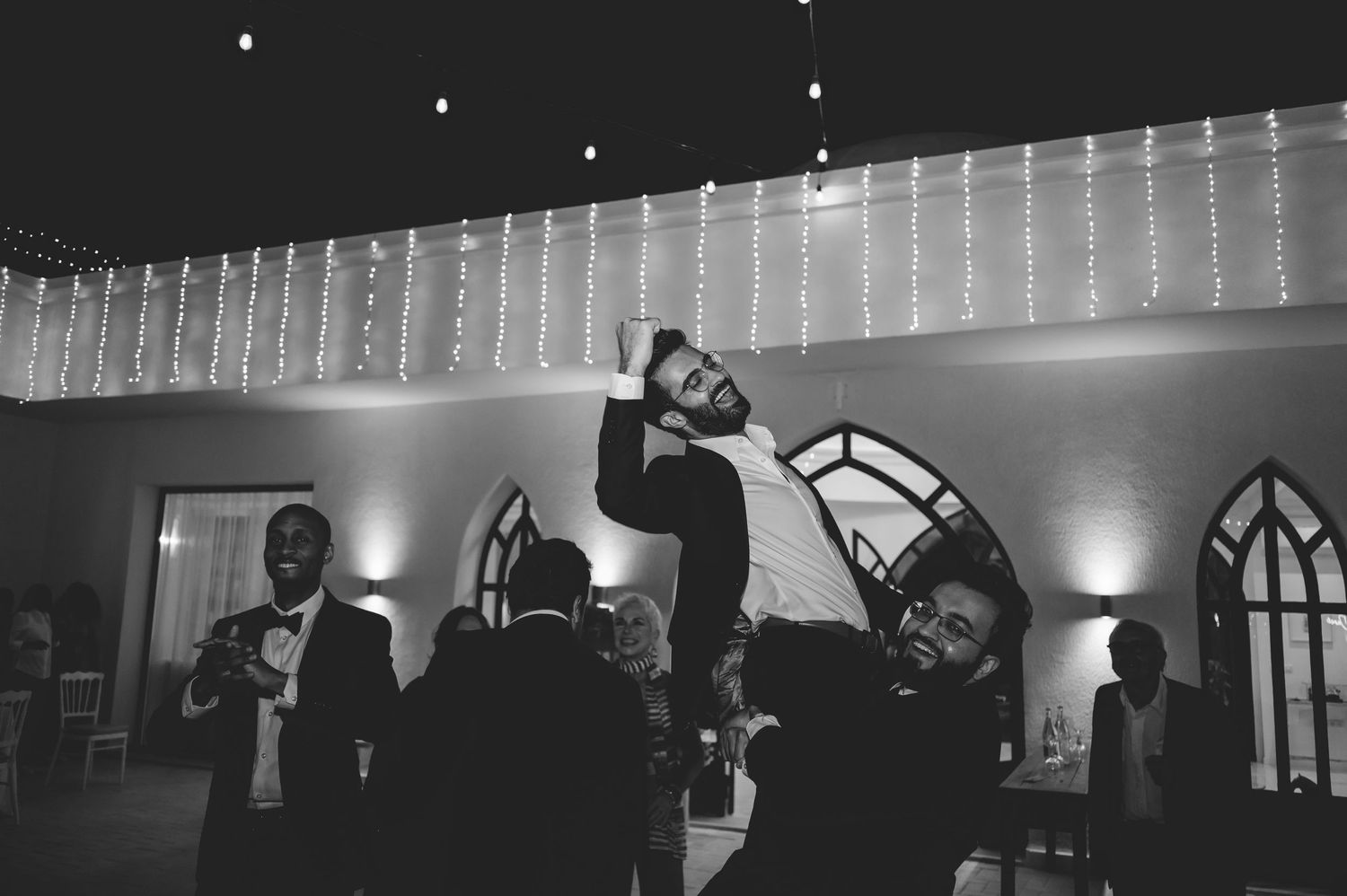 Black and white photo of man dancing energetically with friends at elegant wedding reception.